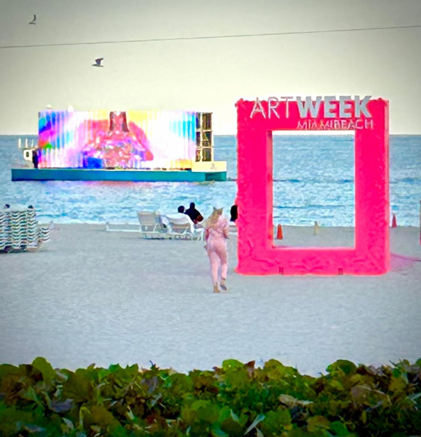 Hot-pink 'Art Week Miami Beach' picture-frame installation on the sand, a floating LED art barge glowing offshore.