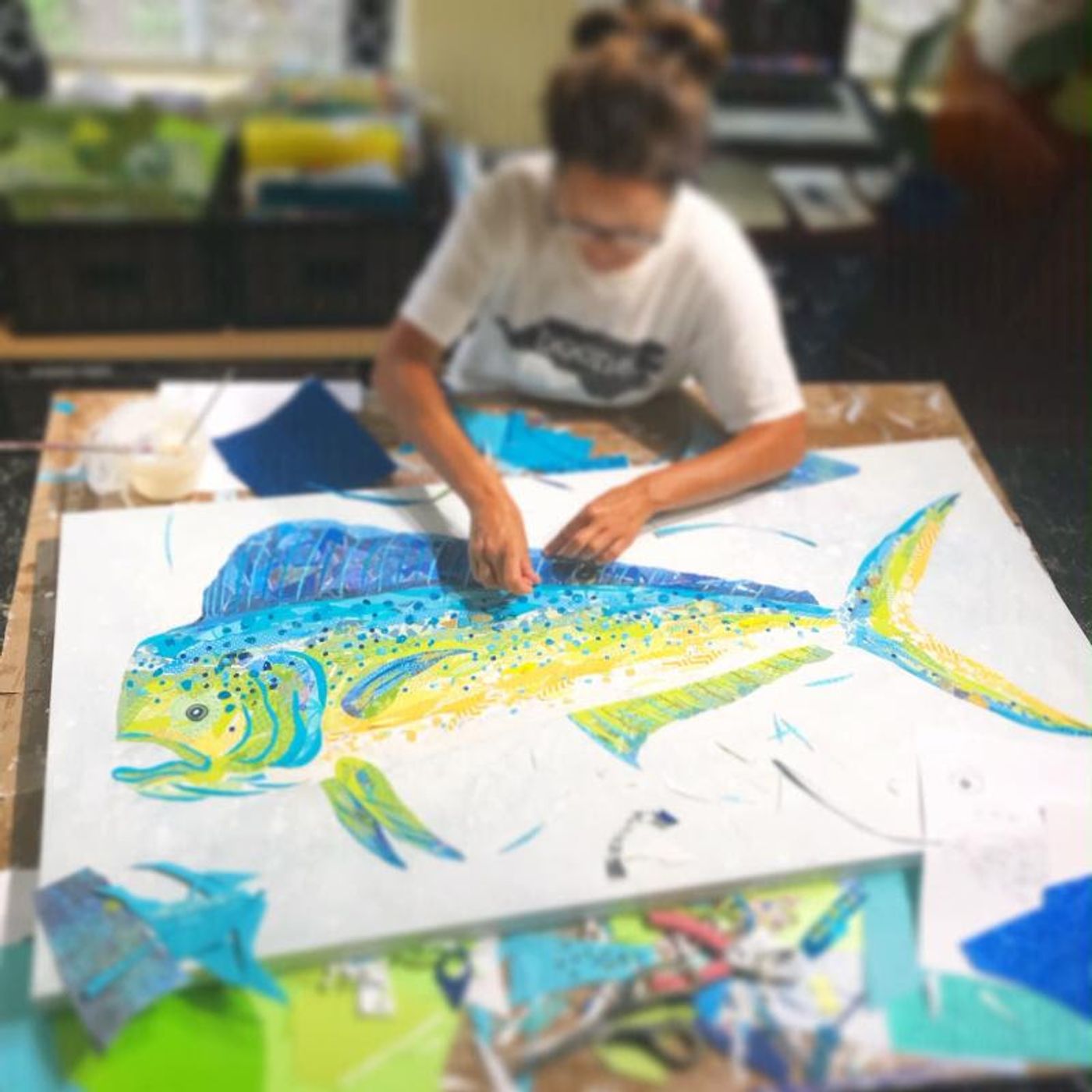Jennifer, hair up and in glasses, pieces a mahi-mahi collage in turquoise, green, and gold papers at her studio table.