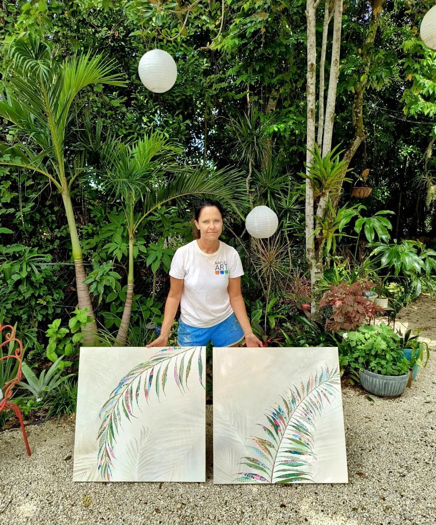 Jennifer stands in her studio holding two new floral collages — layered patterned-paper blooms and leaves in coral, blue, and green.