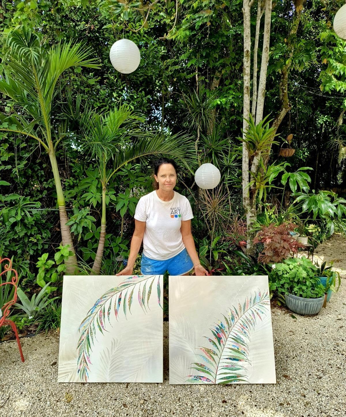 Jennifer stands in her studio holding two new floral collages — layered patterned-paper blooms and leaves in coral, blue, and green.