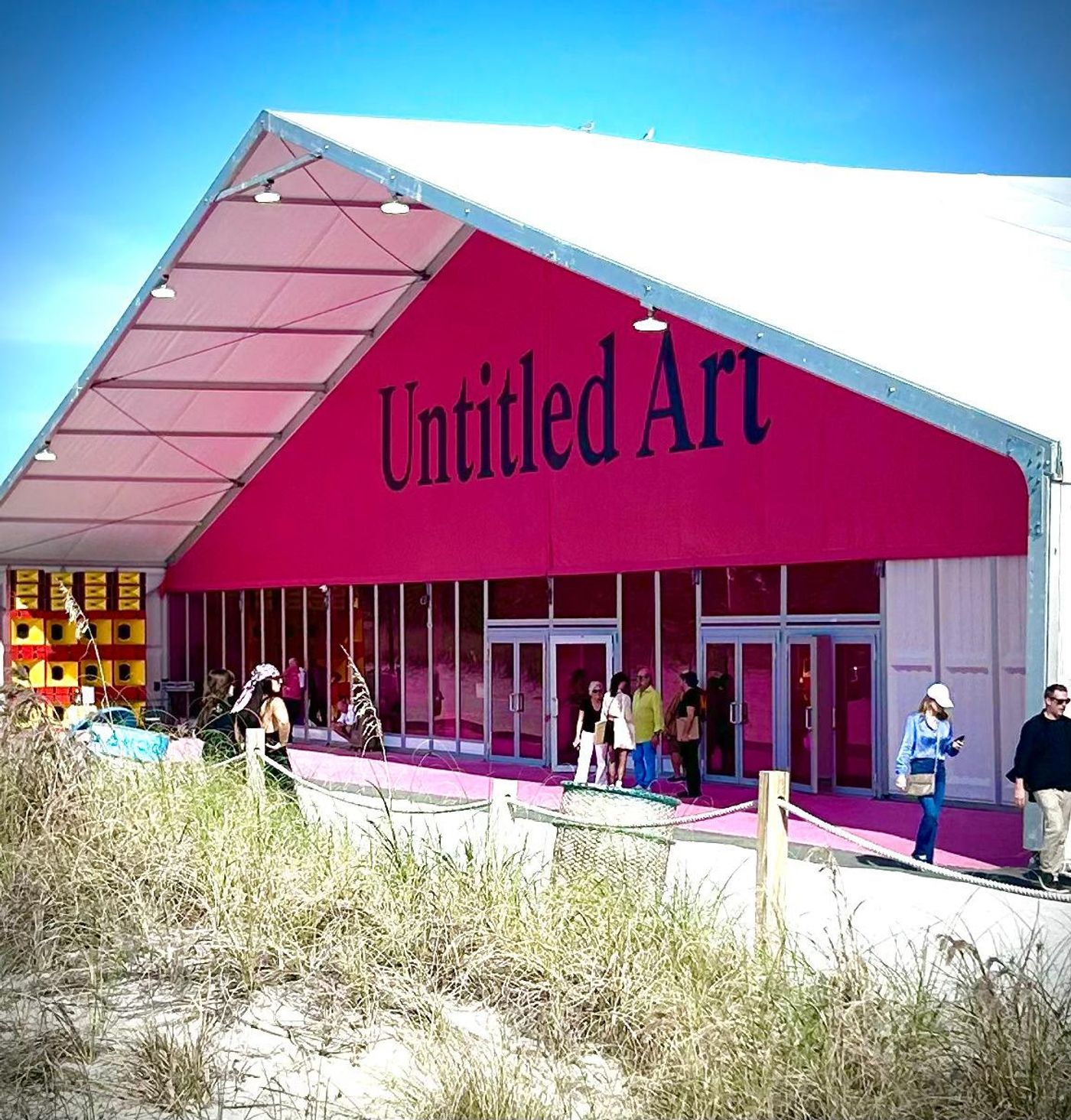 Magenta entrance tent of the Untitled Art fair on Miami Beach, visitors at the glass doors beyond dune grass.