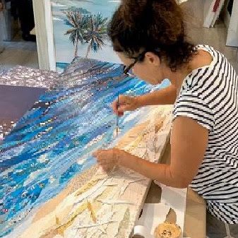 Jennifer detailing the sandy shore of a large ocean collage, rows of tiny blue paper pieces forming the water