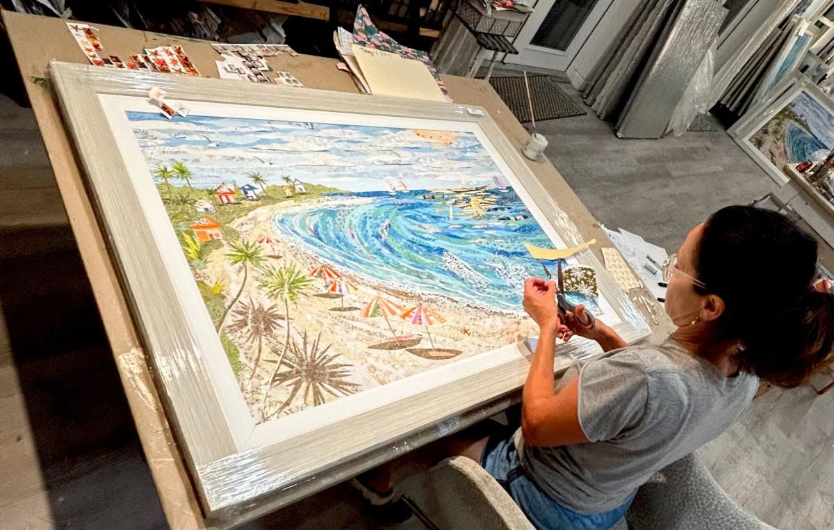 Jennifer cuts paper at her studio worktable, the large framed Sunny Days beach-cove collage laid out before her