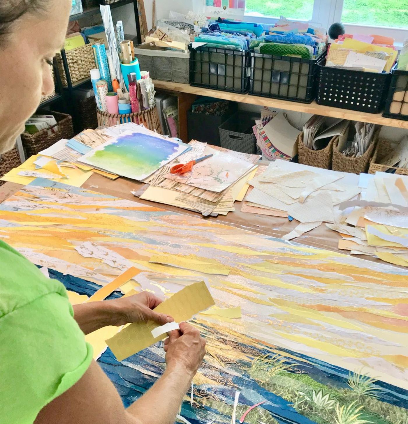 Jennifer peels the backing from a gold honeycomb paper strip for the collage's sunset sky, torn yellow strips covering the table.