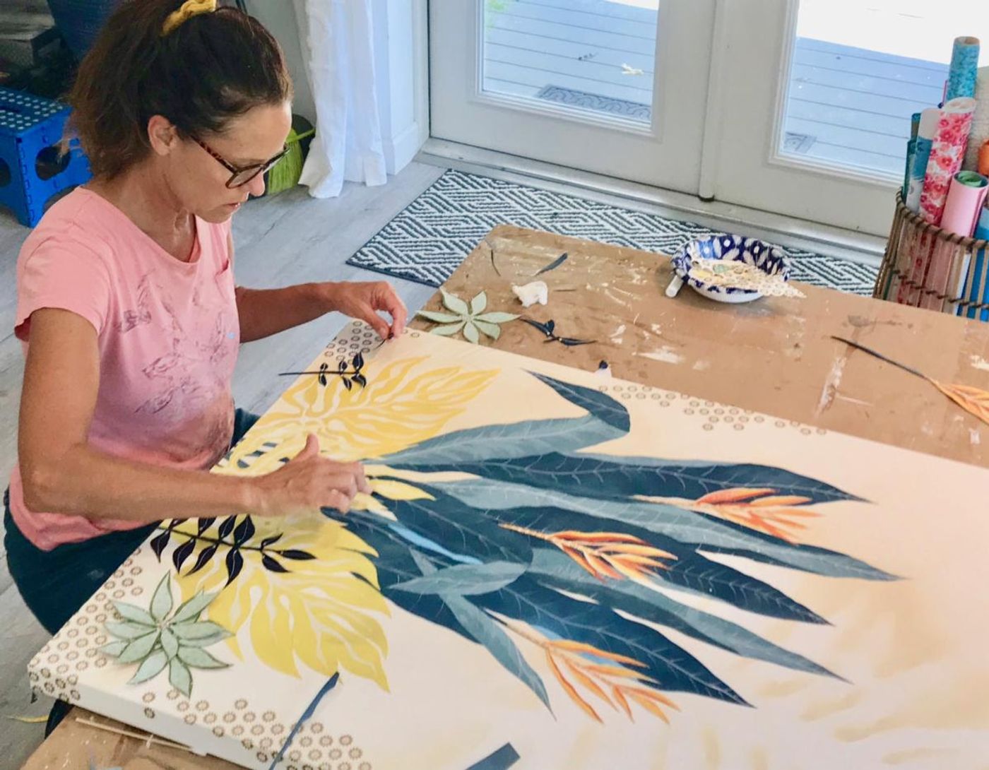 Jennifer arranges hand-cut blue and yellow leaves on a large botanical collage panel in the studio