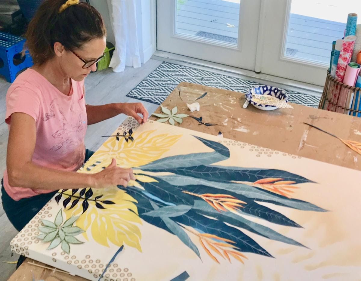 Jennifer arranges hand-cut blue and yellow leaves on a large botanical collage panel in the studio
