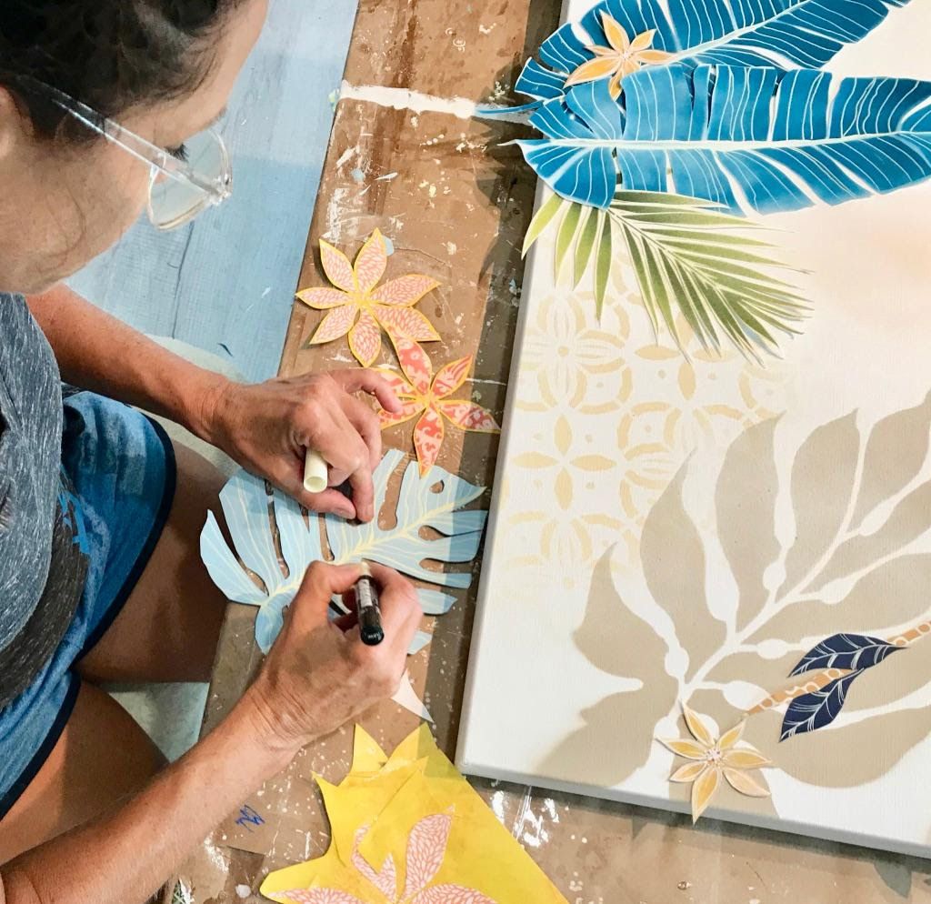Jennifer adds vein lines to a hand-cut pale blue monstera leaf with a marker, beside the in-progress Jungle Love canvas