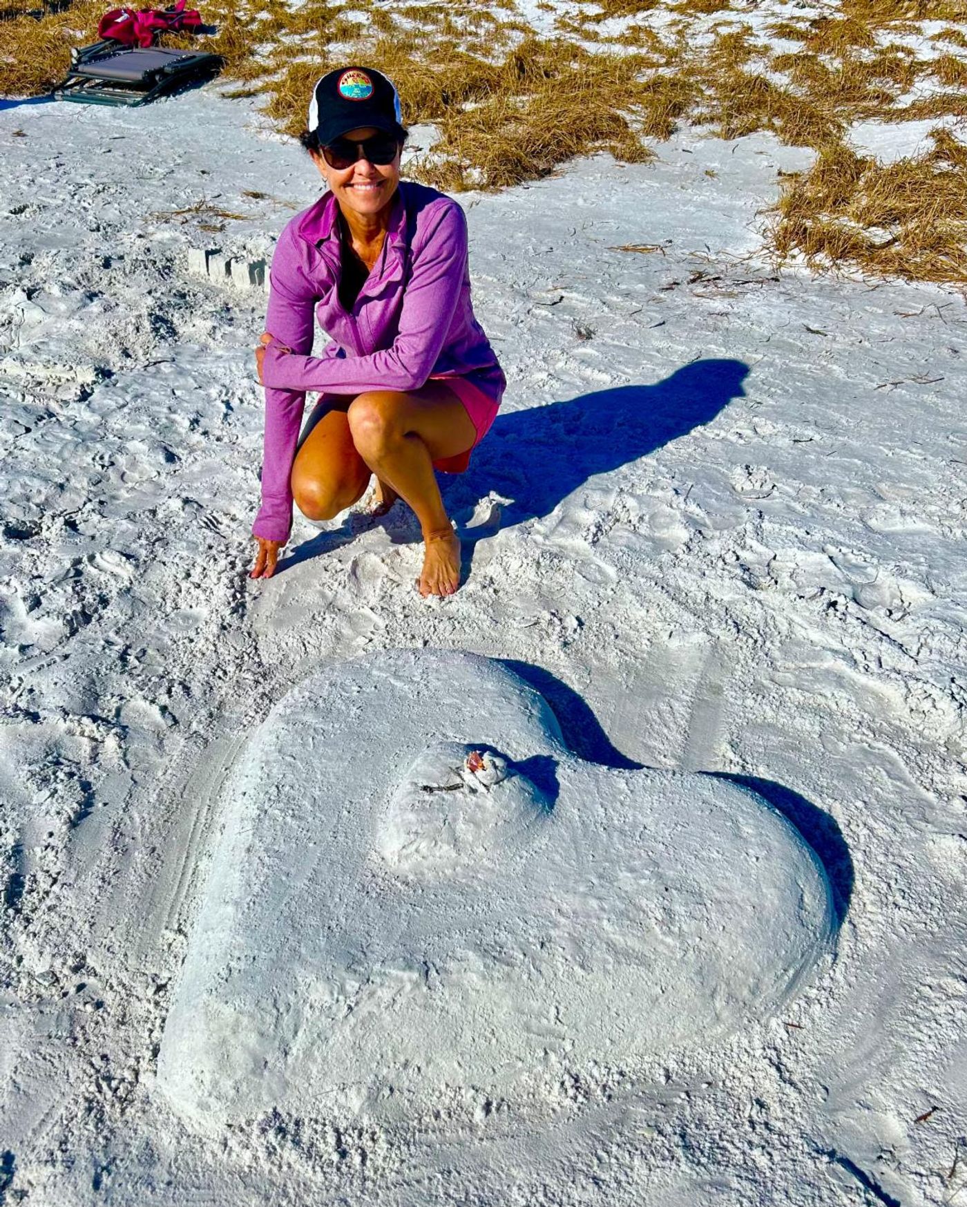 Jennifer crouches barefoot on white sand beside a large heart she sculpted on the beach