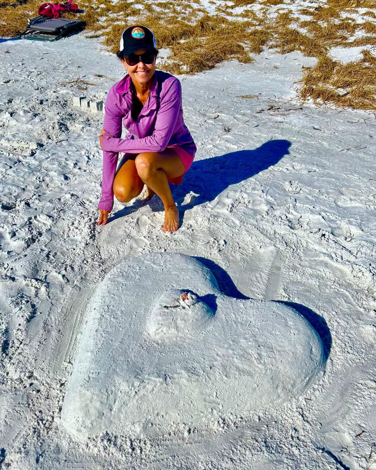 Jennifer crouches barefoot on white sand beside a large heart she sculpted on the beach