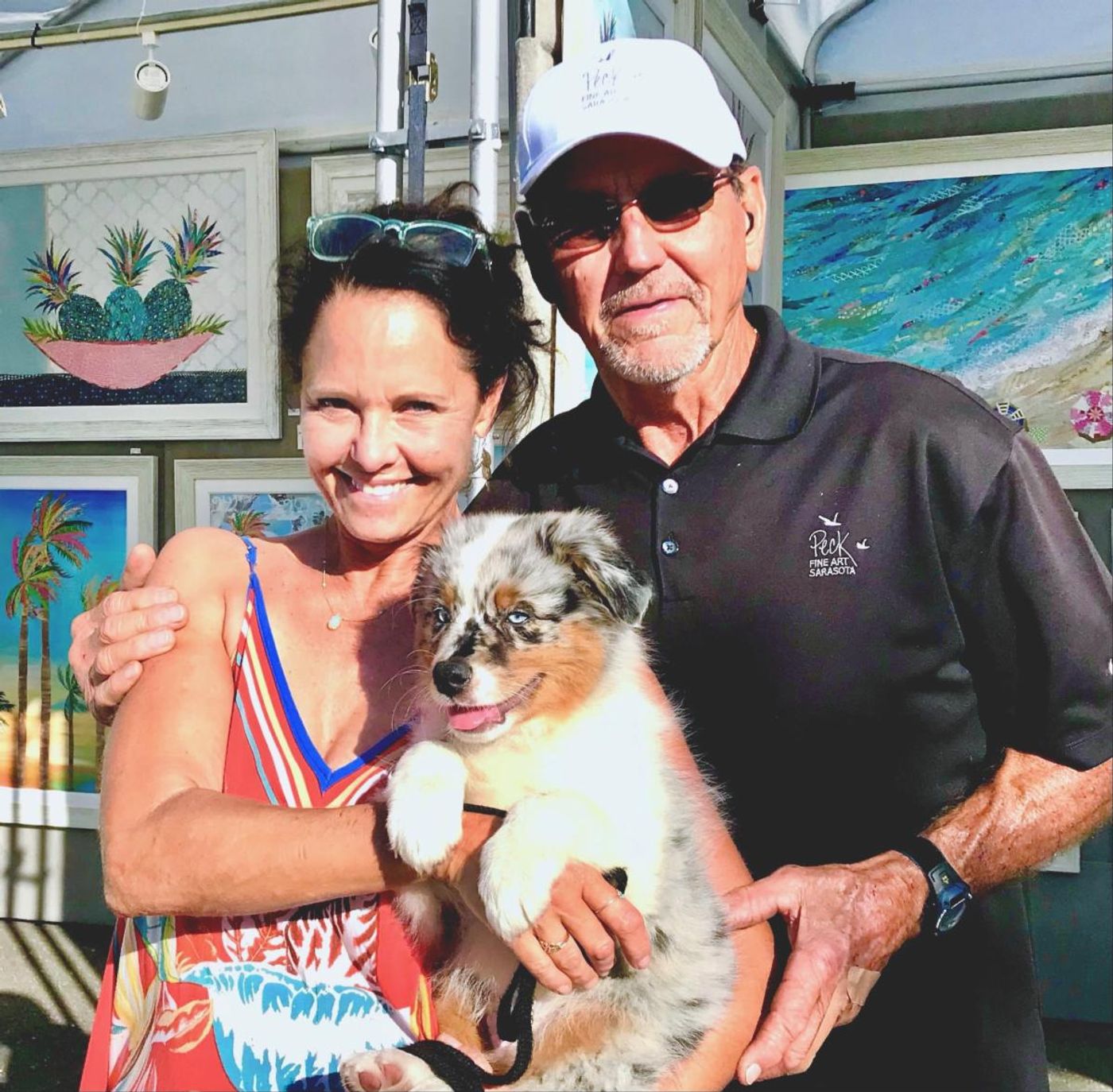 Jennifer and a man in a Peck Fine Art Sarasota polo cradling a blue-merle puppy in the booth, coastal collages behind them