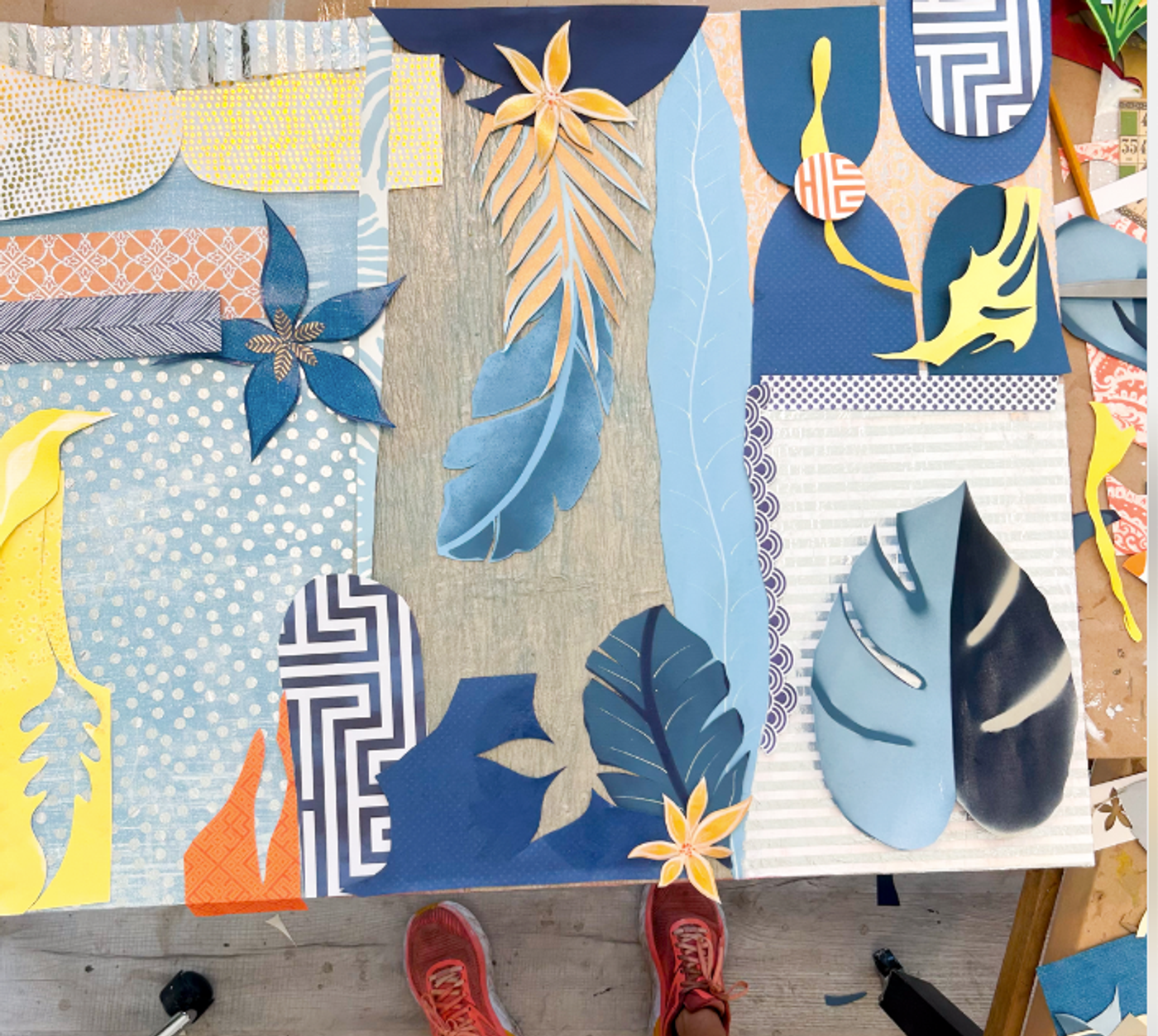 Looking down at a large leaf collage in progress, blue fronds, peach star flowers and patterned papers, red sneakers at the edge