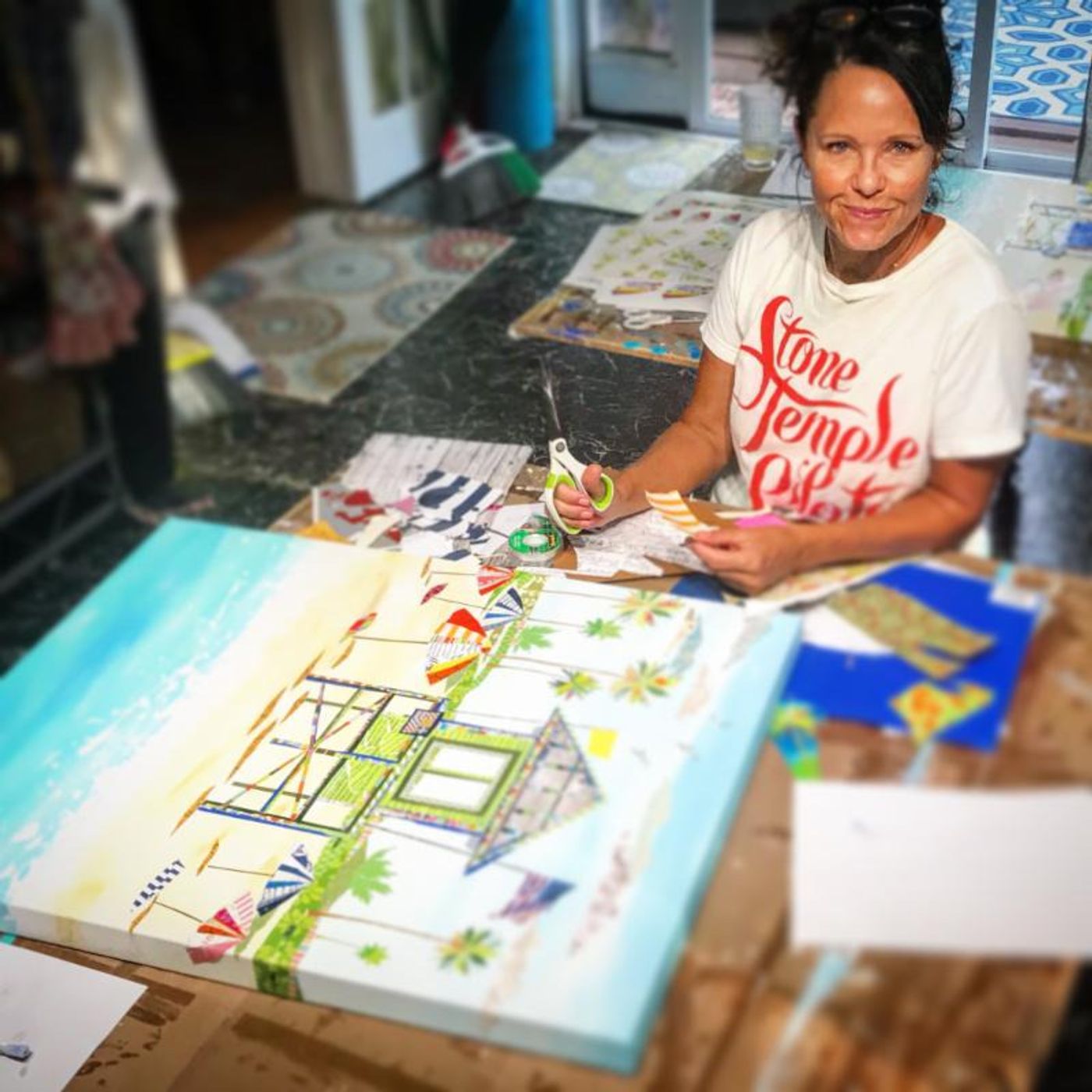 Jennifer smiling at the worktable, scissors in hand, a beach-hut collage with a turquoise sky in front of her
