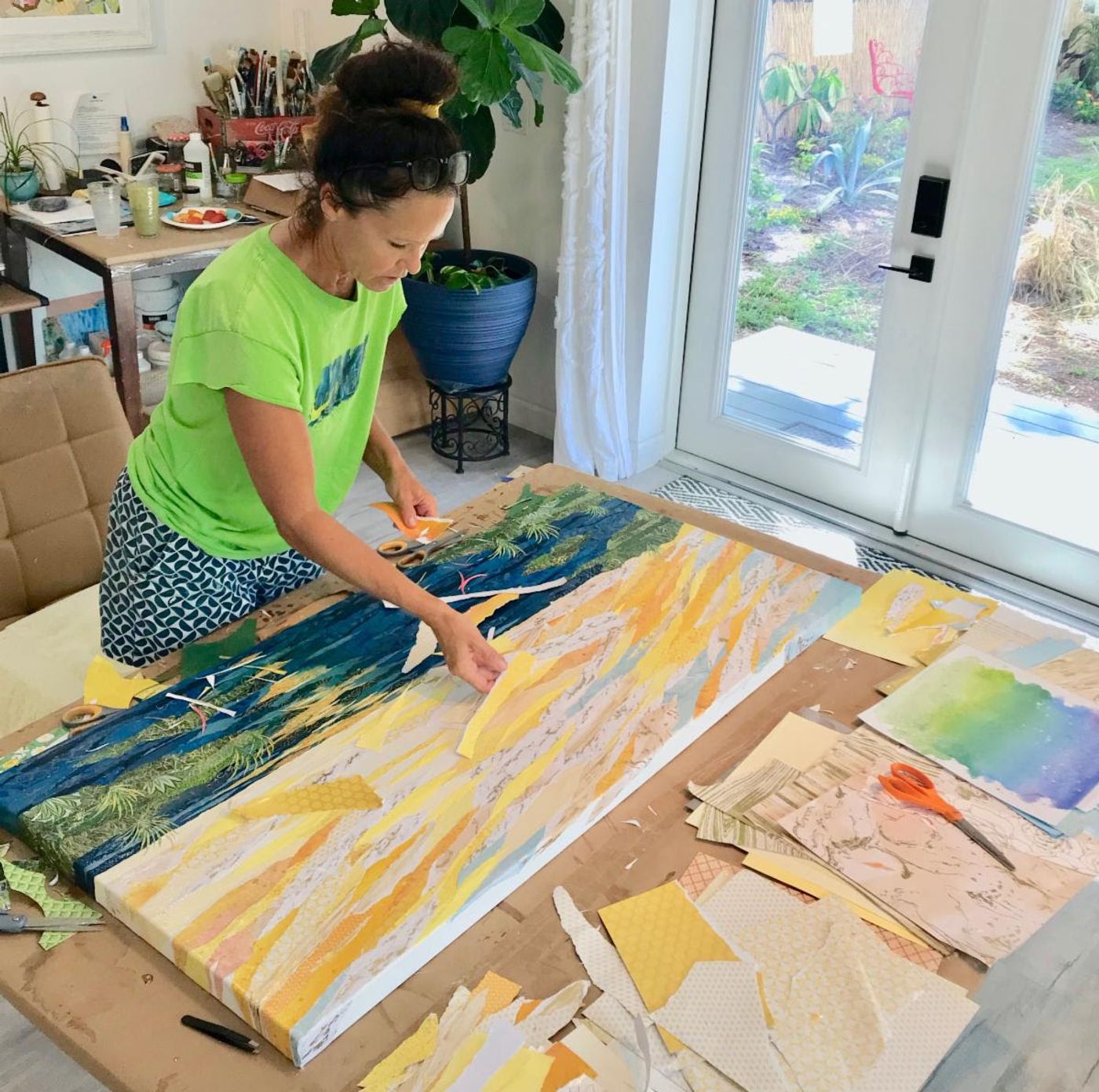 Jennifer leans over her studio work table, laying golden paper strips onto the wide bay-at-sunset collage in progress.
