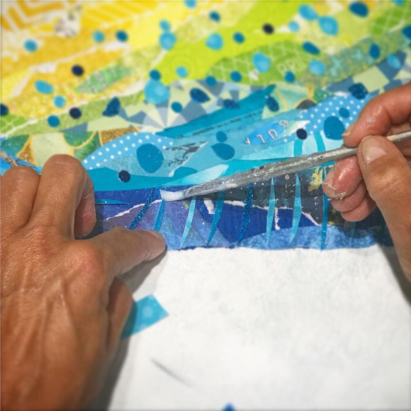 Close-up of Jennifer's hands gluing slim blue paper curves into a wave with a silver tool, yellow-green layers above.