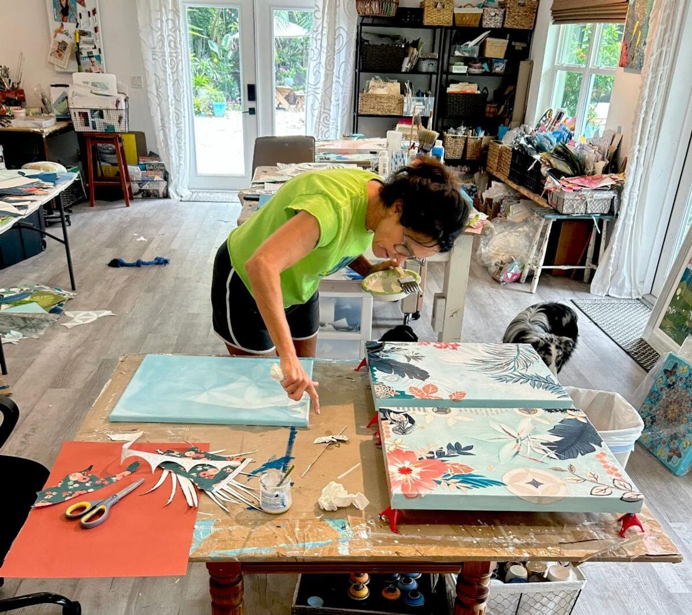 Jennifer at her studio worktable starting a pale aqua canvas beside two tropical collages in progress, her dog nearby
