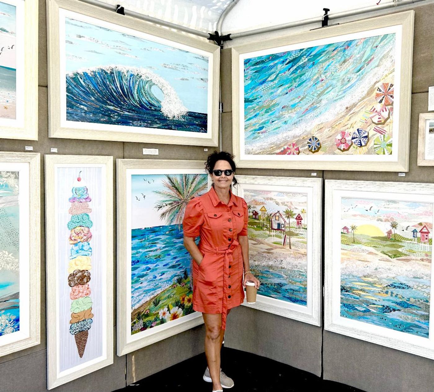 Jennifer stands in her festival booth among framed coastal collages — a cresting wave, beach umbrellas from above, an ice cream cone stack