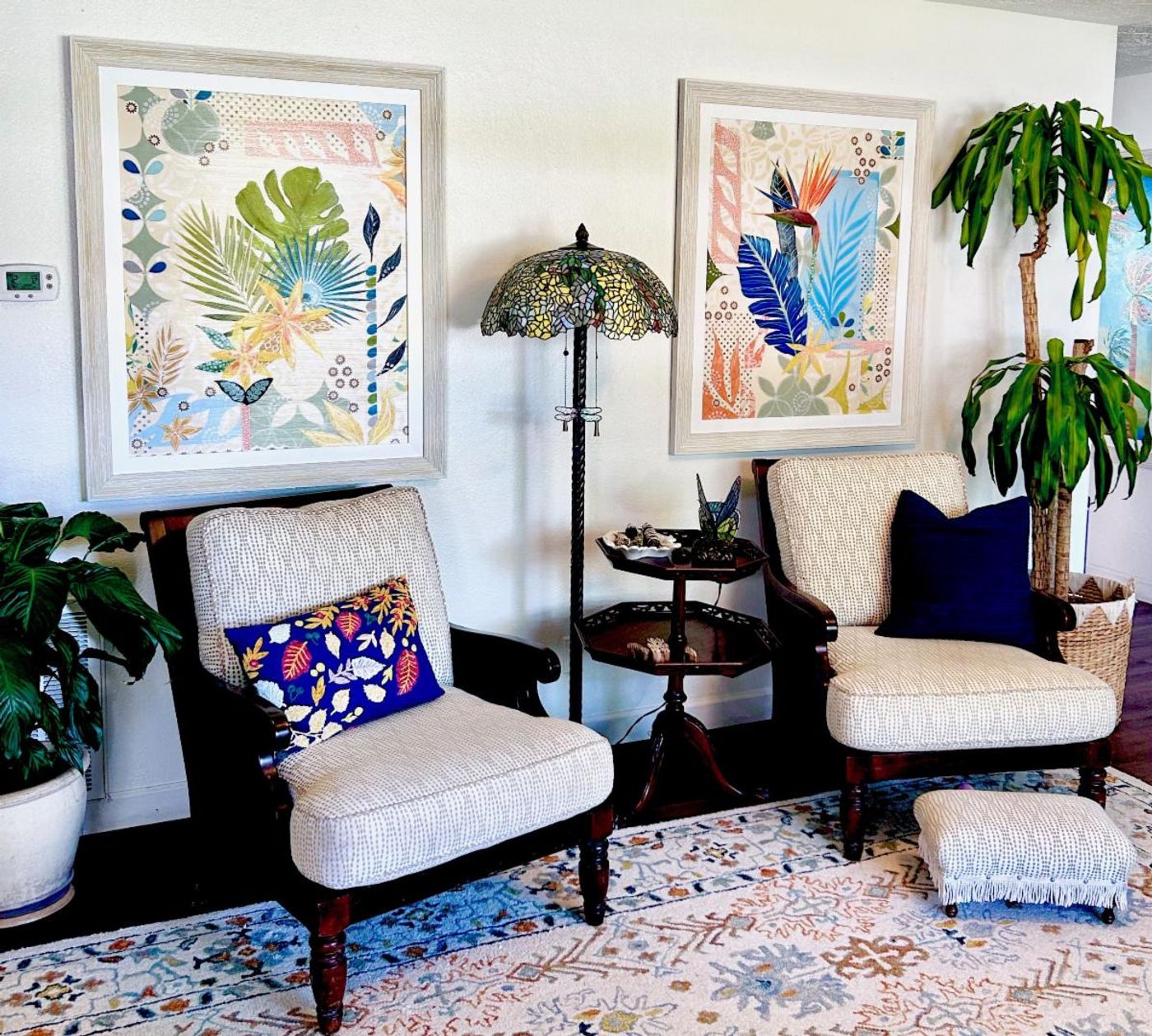 Two large framed botanical collages above a pair of armchairs in a light-filled sitting room with a stained-glass lamp