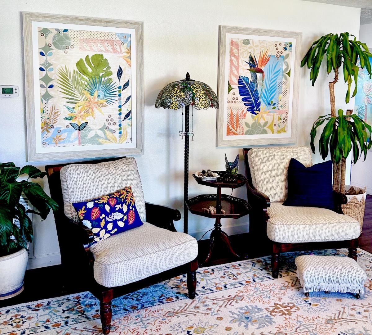 Two large framed botanical collages above a pair of armchairs in a light-filled sitting room with a stained-glass lamp