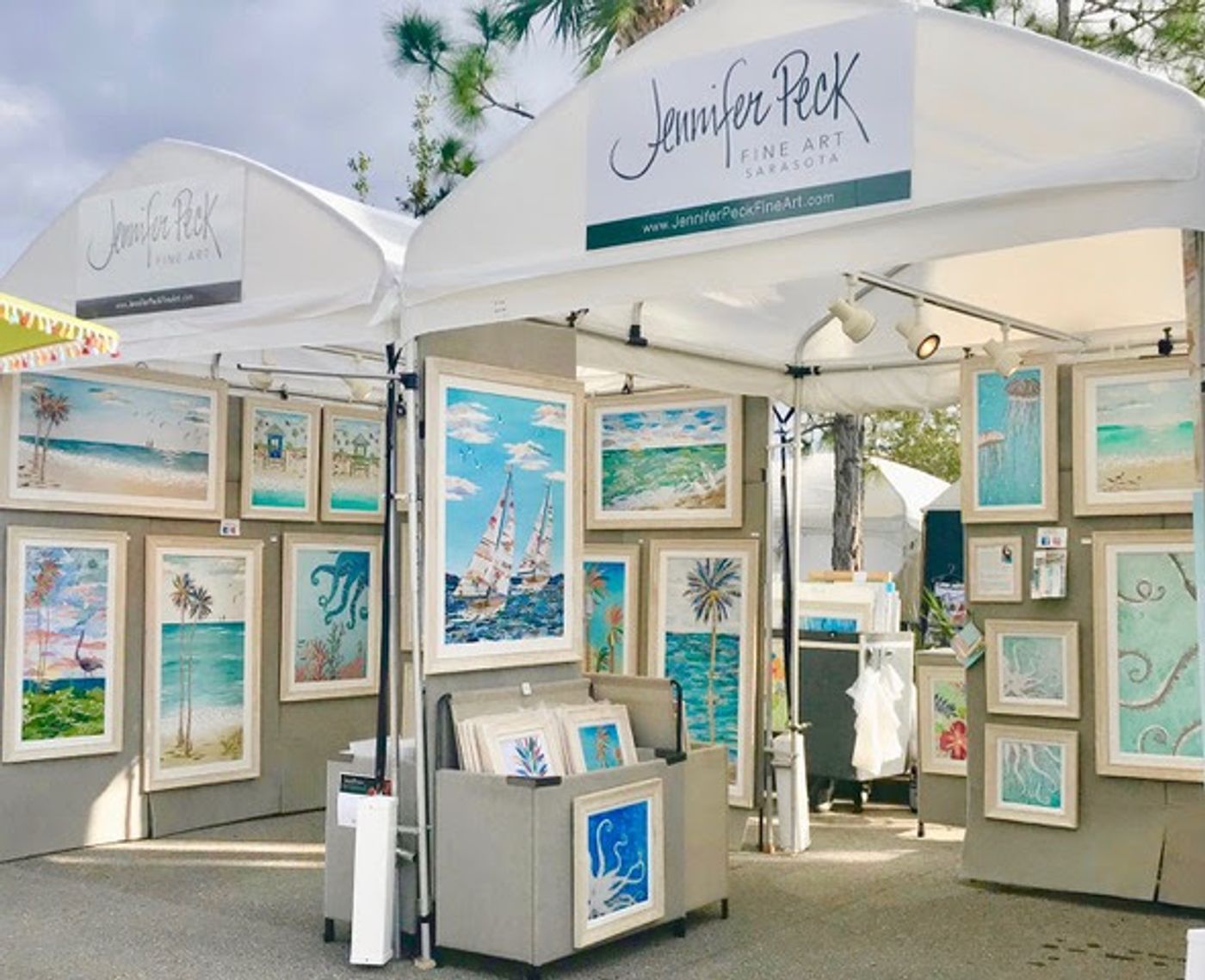 Two white festival tents with Jennifer Peck Fine Art banners, framed coastal collages covering the walls, a print bin at the center.