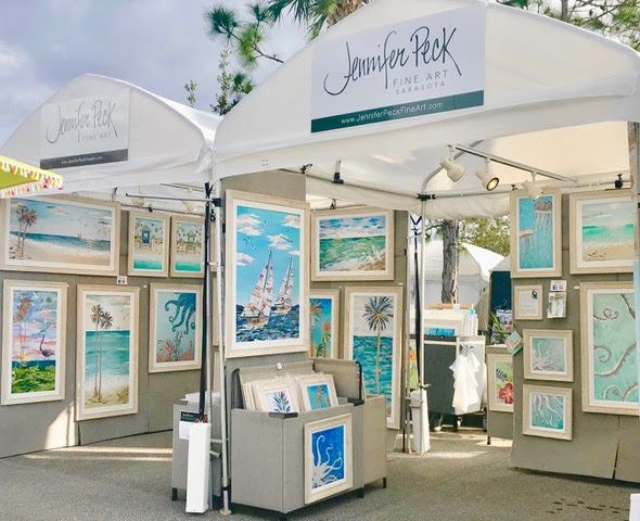 Two white festival tents with Jennifer Peck Fine Art banners, framed coastal collages covering the walls, a print bin at the center.