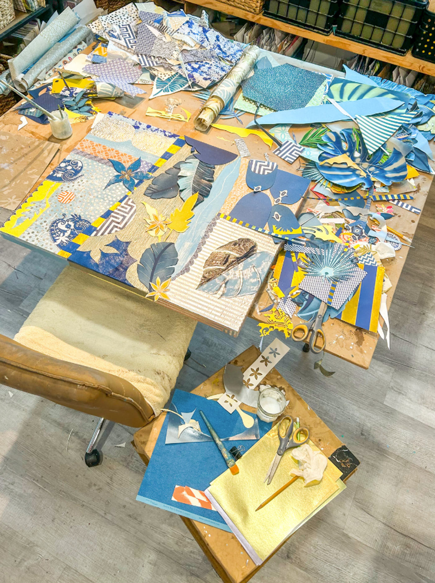 Overhead view of the studio table, a scrap collage taking shape amid piles of blue patterned paper, scissors and gold sheets