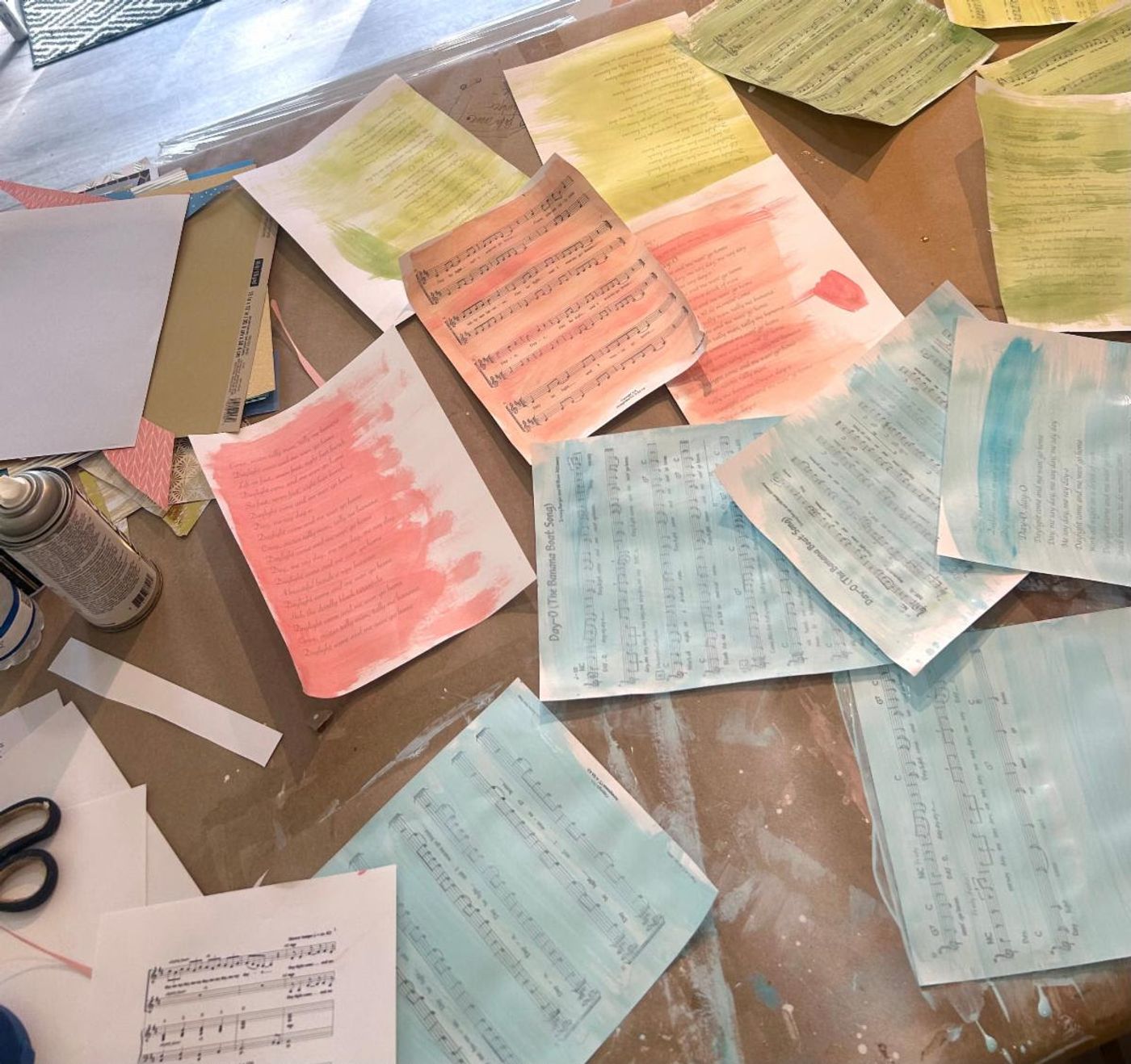Day-O sheet music and lyric pages brushed with coral, aqua, and green washes, drying across the studio worktable