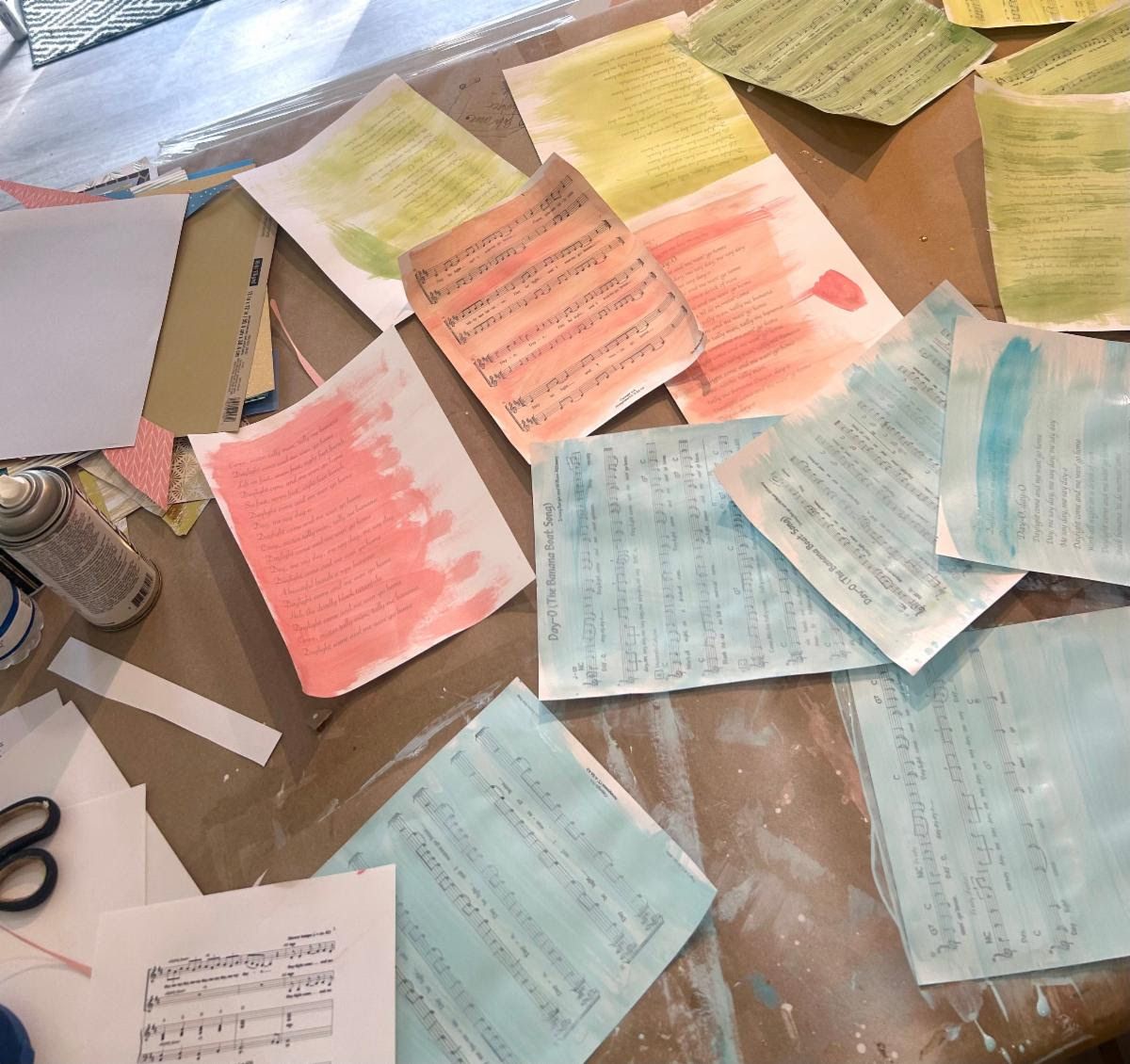 Day-O sheet music and lyric pages brushed with coral, aqua, and green washes, drying across the studio worktable