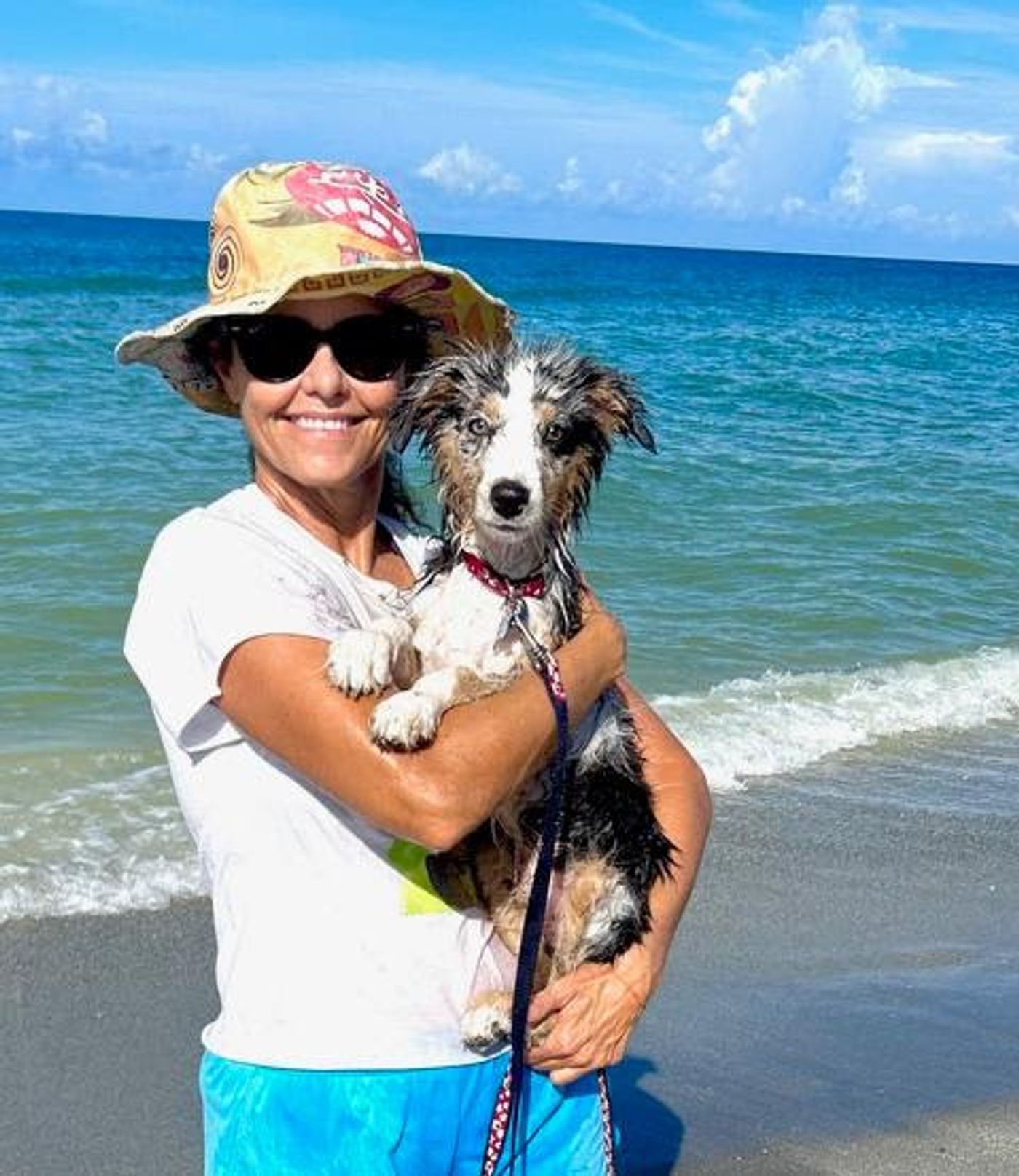 Jennifer in a sun hat and sunglasses holds the wet puppy at the water's edge, turquoise waves rolling in behind them.