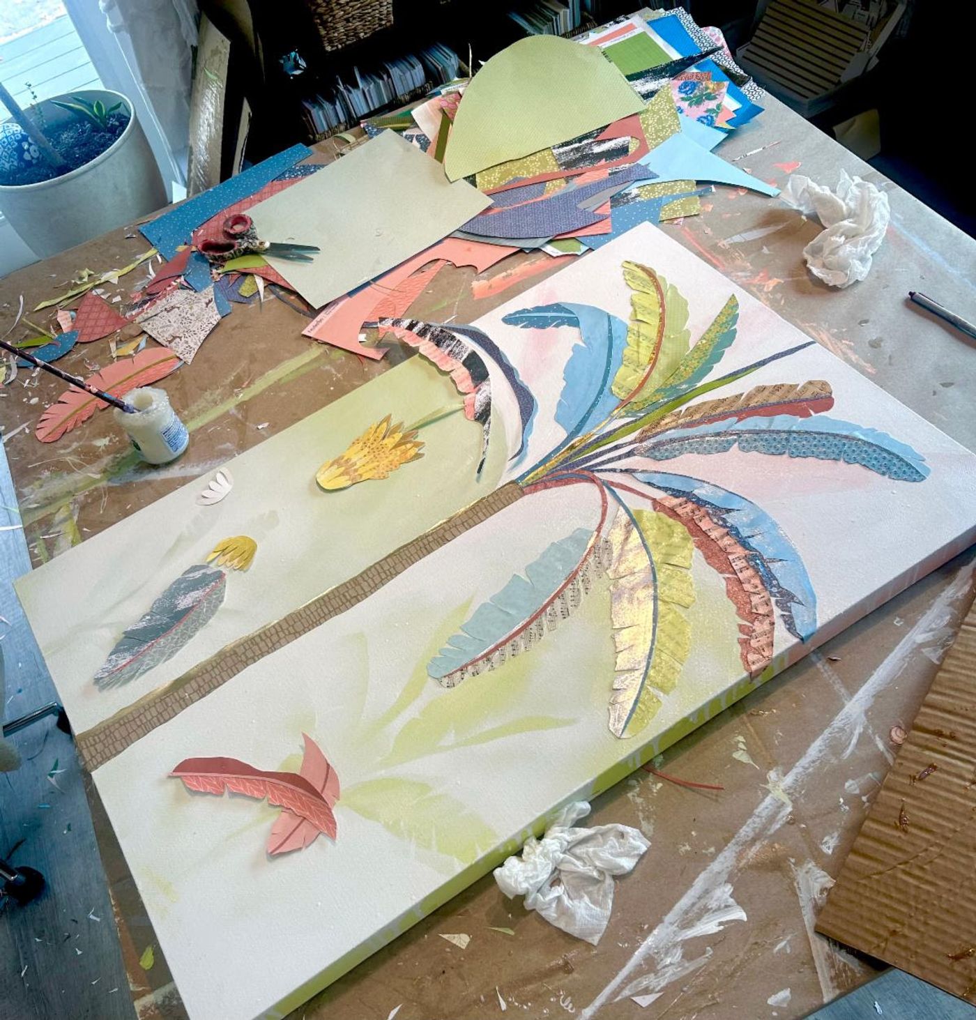 Two tall banana palm panels in progress, cut-paper leaves and patterned trunks laid over soft green washes on the worktable