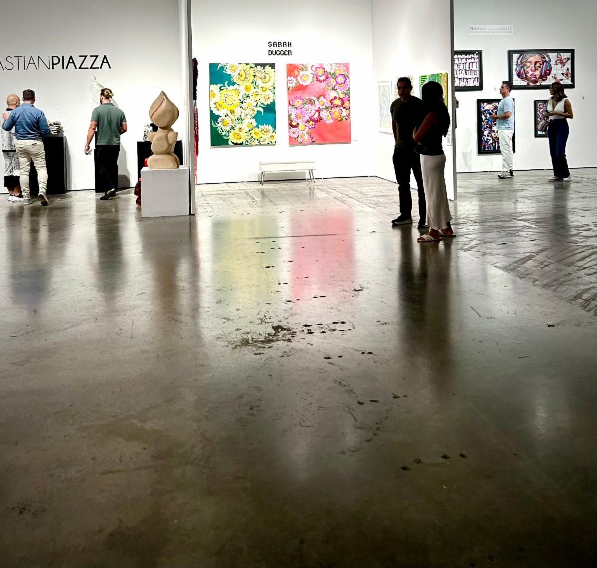 Fairgoers wander a white-walled Miami hall between Sarah Dugger's floral canvases and a carved wooden sculpture.