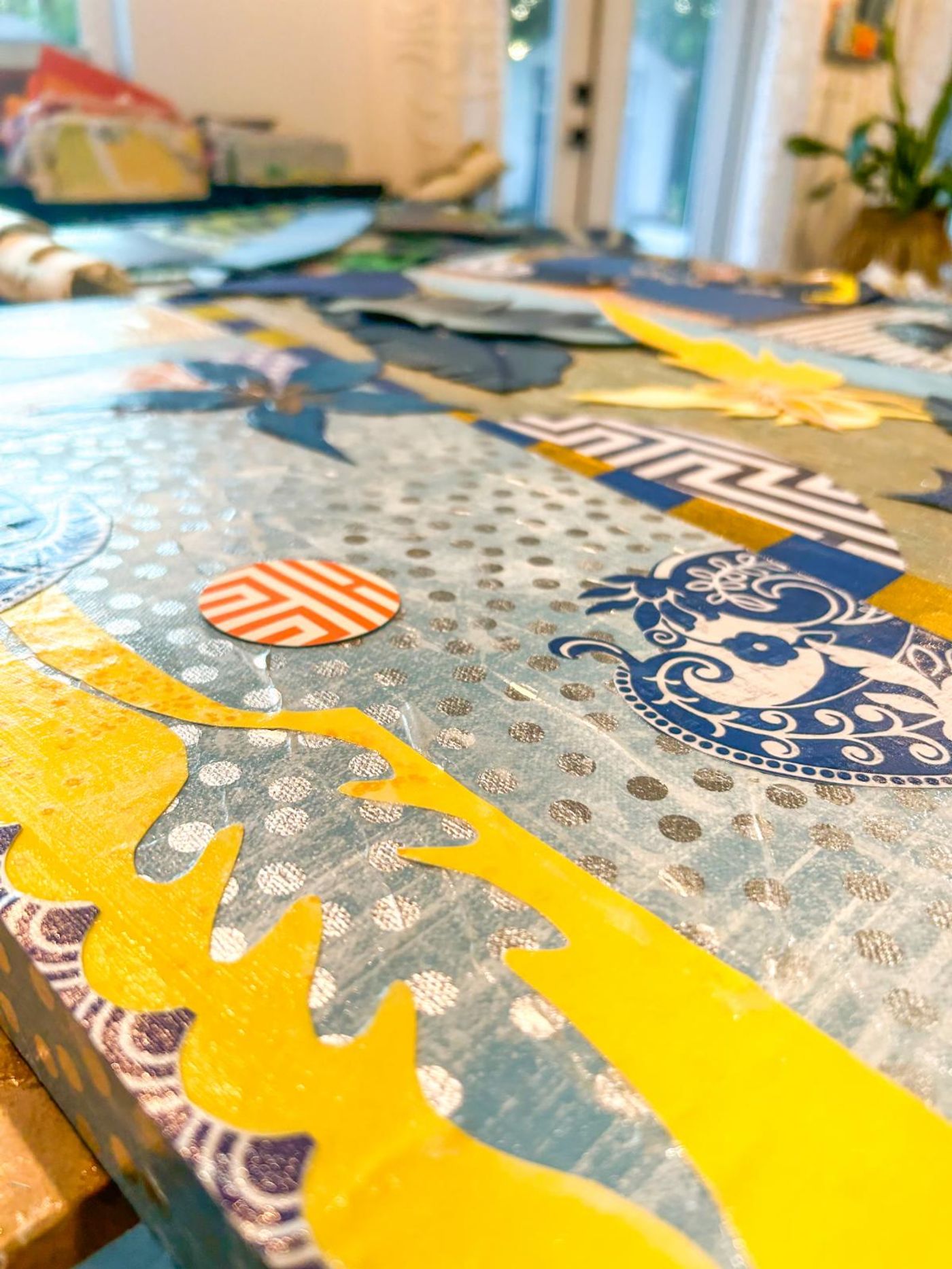 Close-up across a sealed collage panel: yellow cut shapes, silver dots and a blue-and-white china motif catching the light