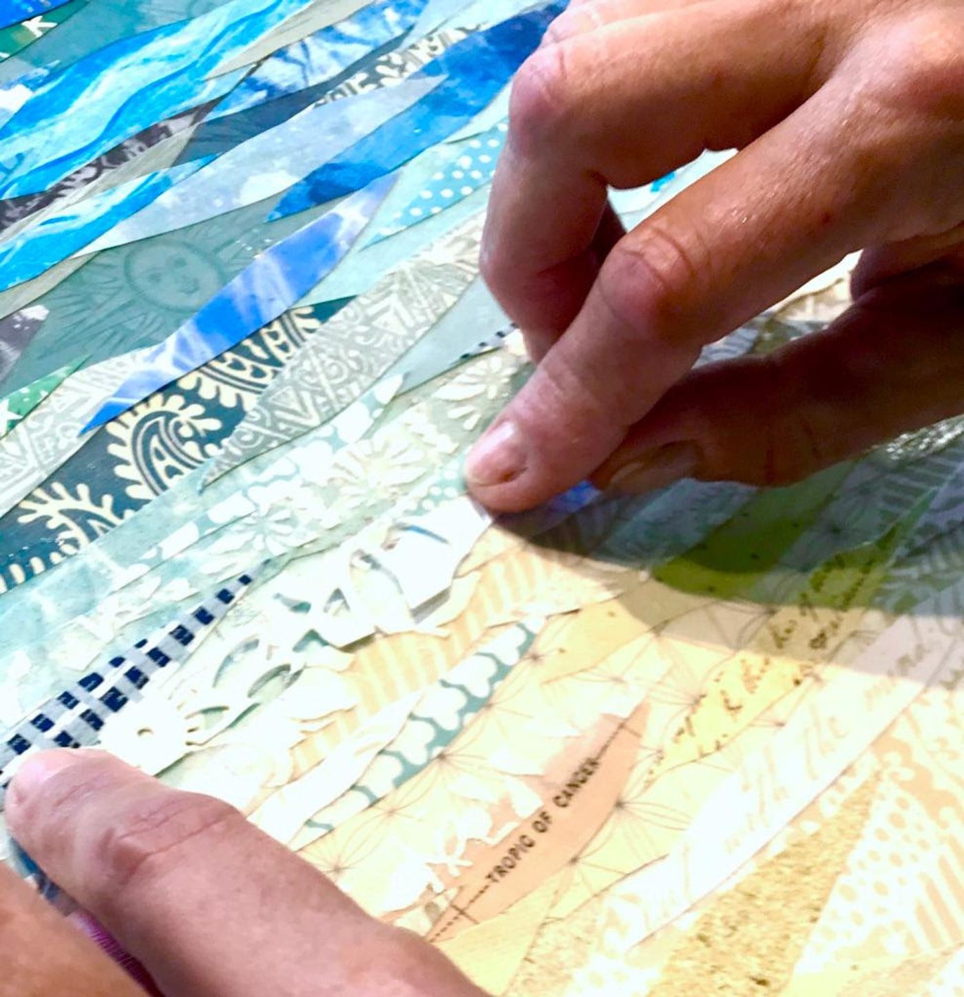 Jennifer at her studio worktable placing cut paper on a pale blue canvas, tropical collages in progress and her dog looking on