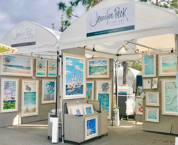 Two white festival tents with Jennifer Peck Fine Art banners, walls of framed coastal collages and a browsing bin of prints