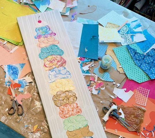 The ice cream cone canvas mid-build on the studio table, surrounded by colorful cut paper scraps and two pairs of scissors.
