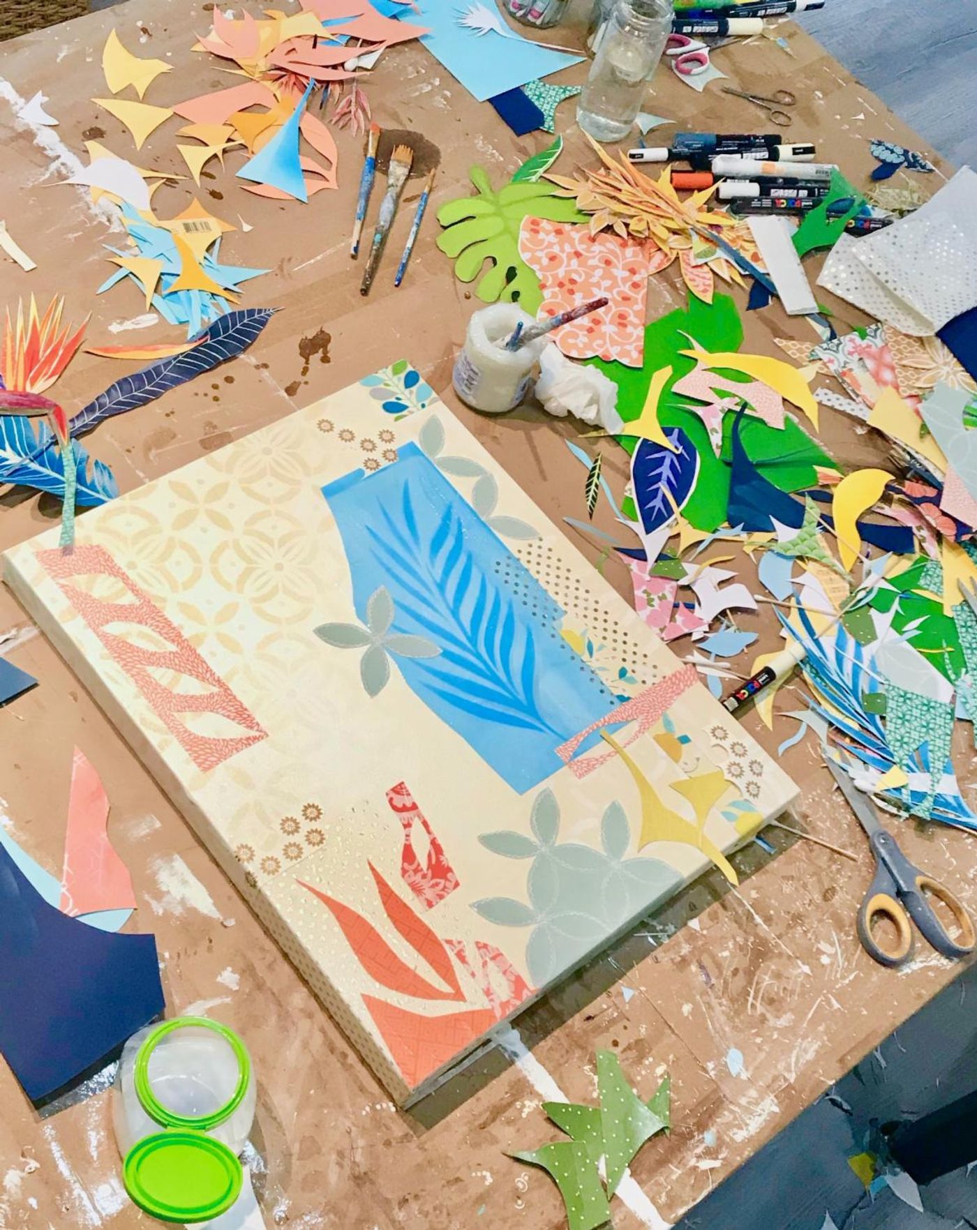 Abstract botanical collage in progress — a blue palm frond on warm cream — on a worktable covered in cut-paper scraps and brushes