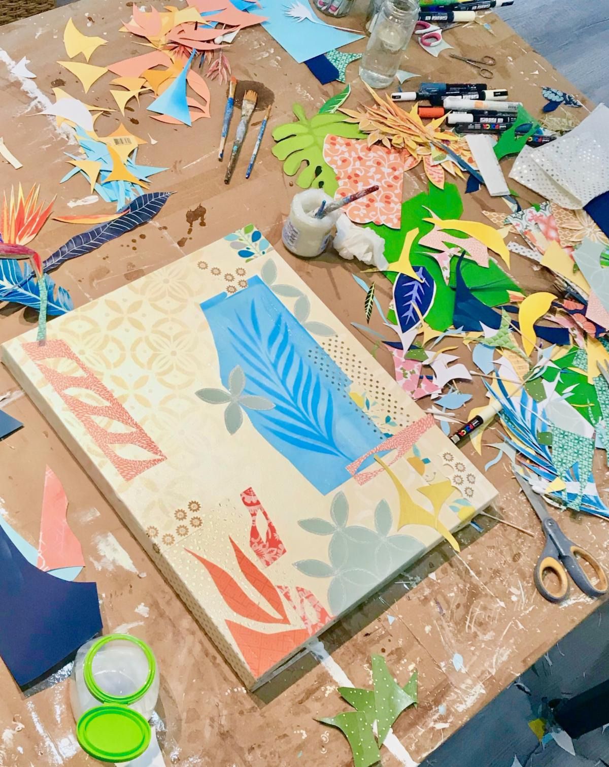 Abstract botanical collage in progress — a blue palm frond on warm cream — on a worktable covered in cut-paper scraps and brushes