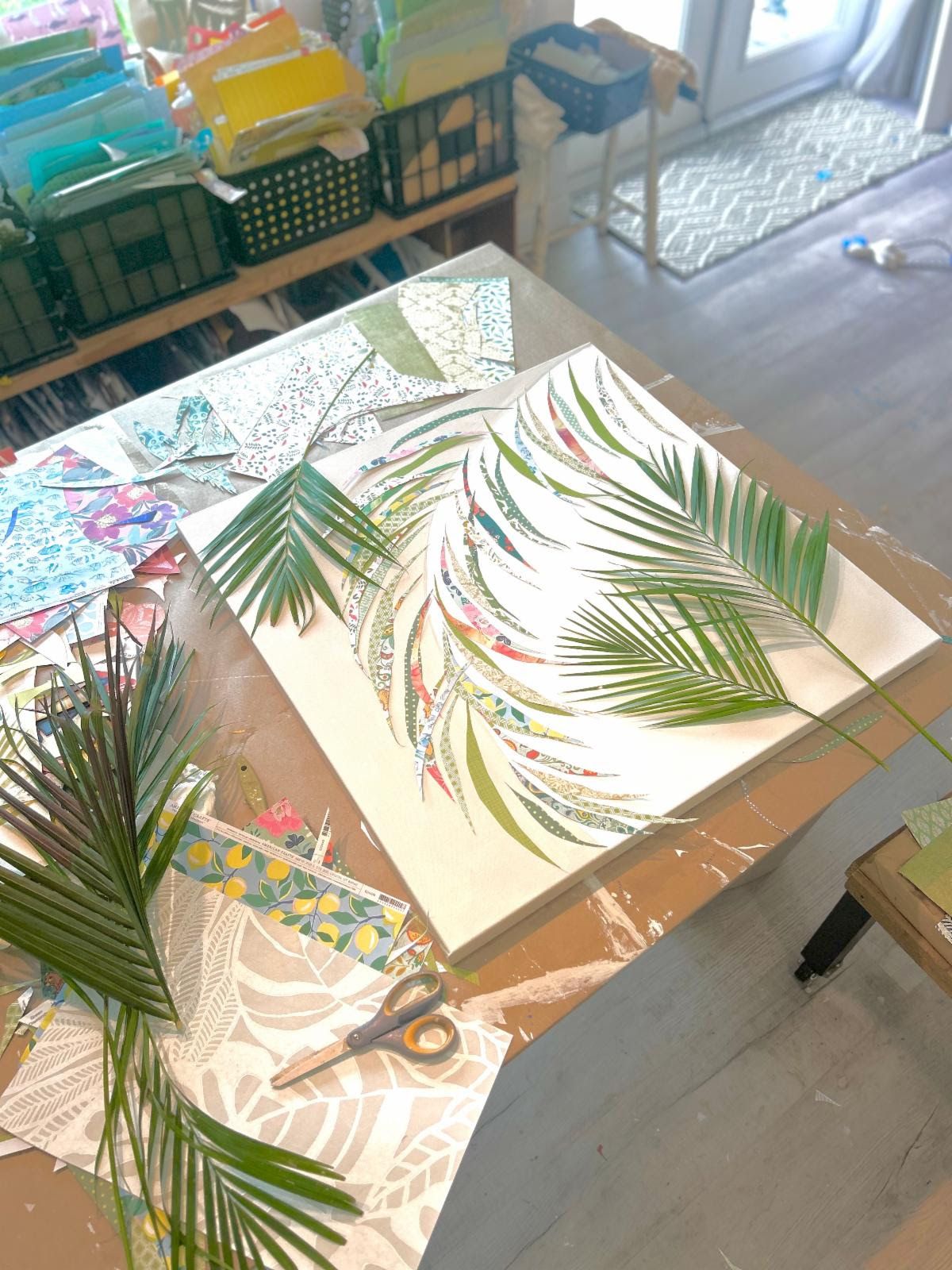 Studio work table with a palm collage in progress — real palm fronds laid on the canvas beside leaflets cut from patterned papers.