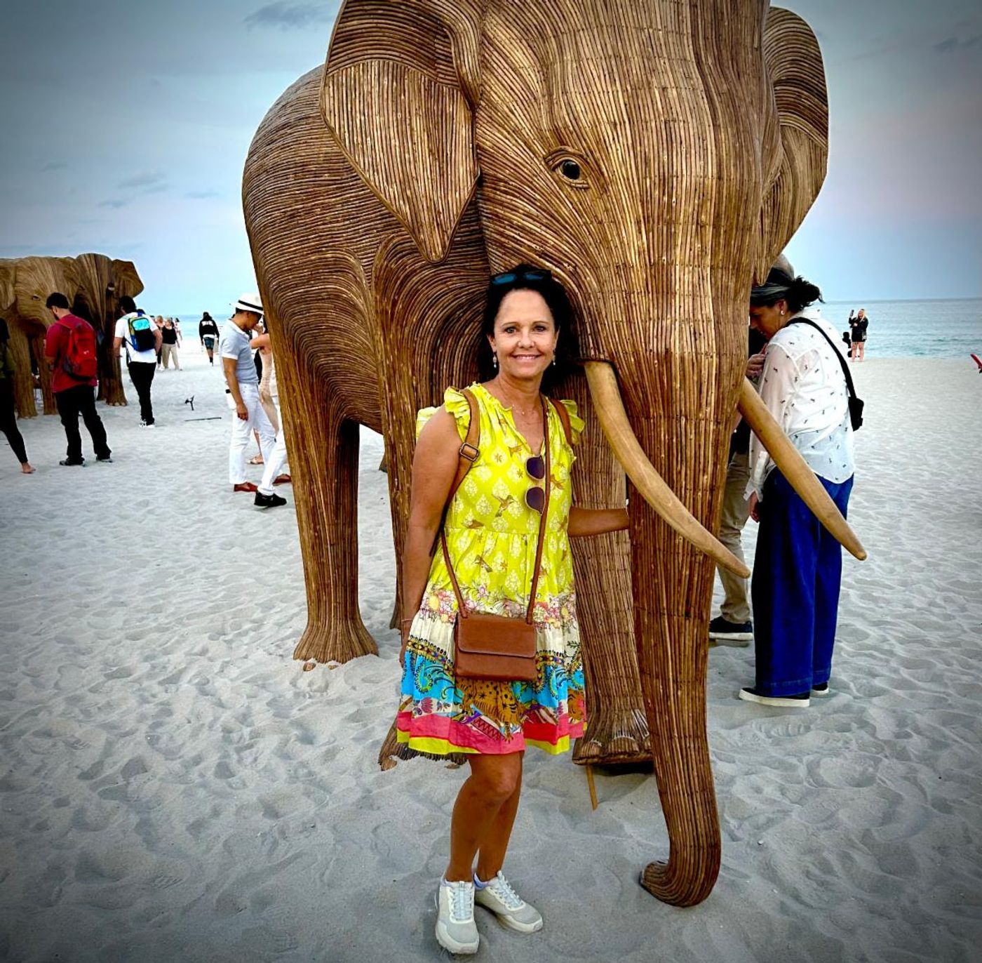 Jennifer in a yellow dress smiles beside a life-size rattan elephant sculpture on the Miami Beach sand, ocean behind.