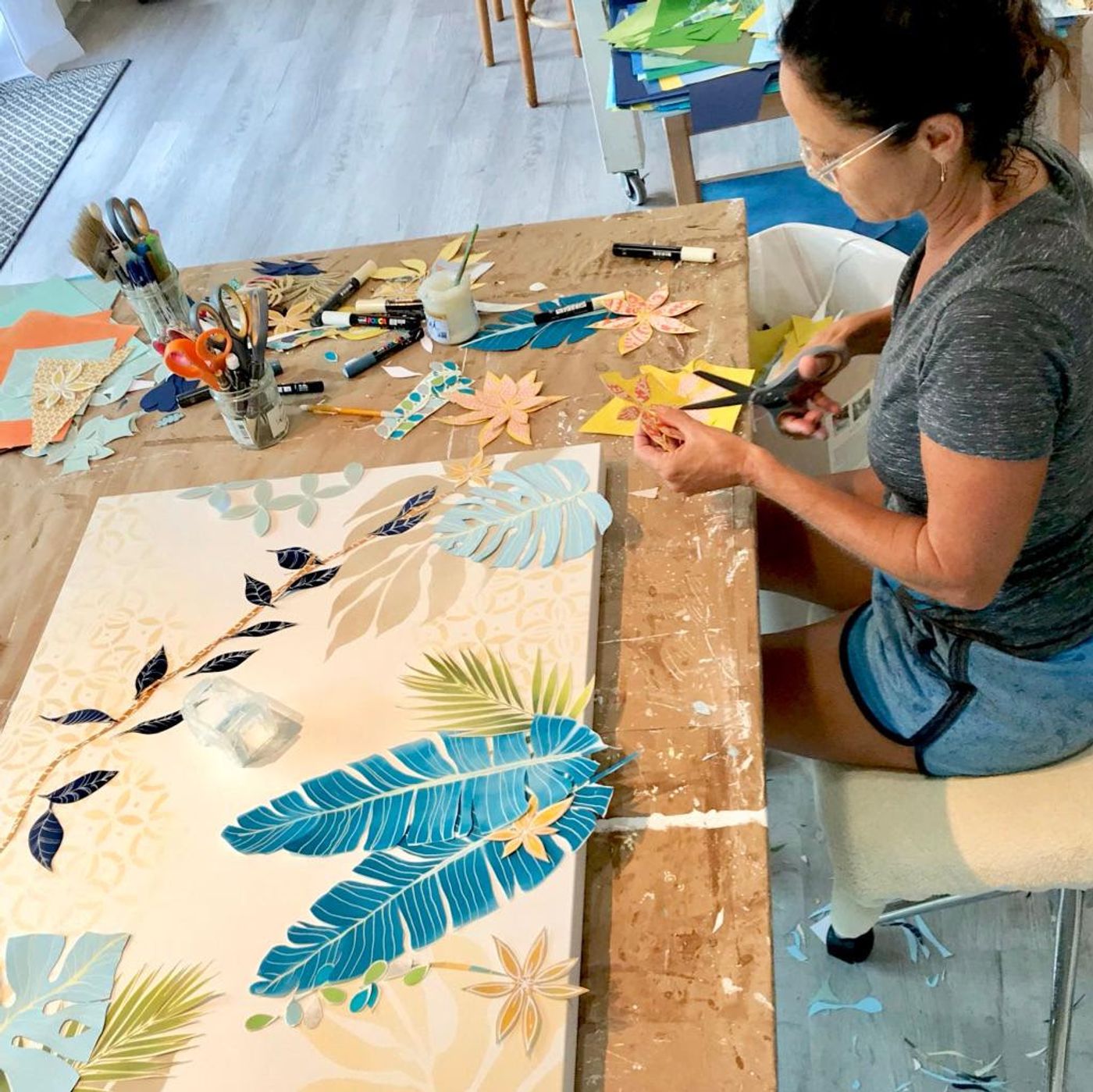 Jennifer cuts paper petals at the worktable while a jungle collage takes shape with blue banana leaves and palm fronds