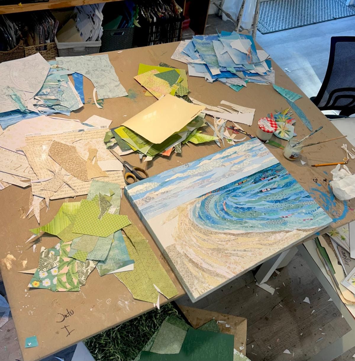 Studio worktable strewn with cut paper scraps and scissors as the Sunny Days shoreline takes shape on canvas