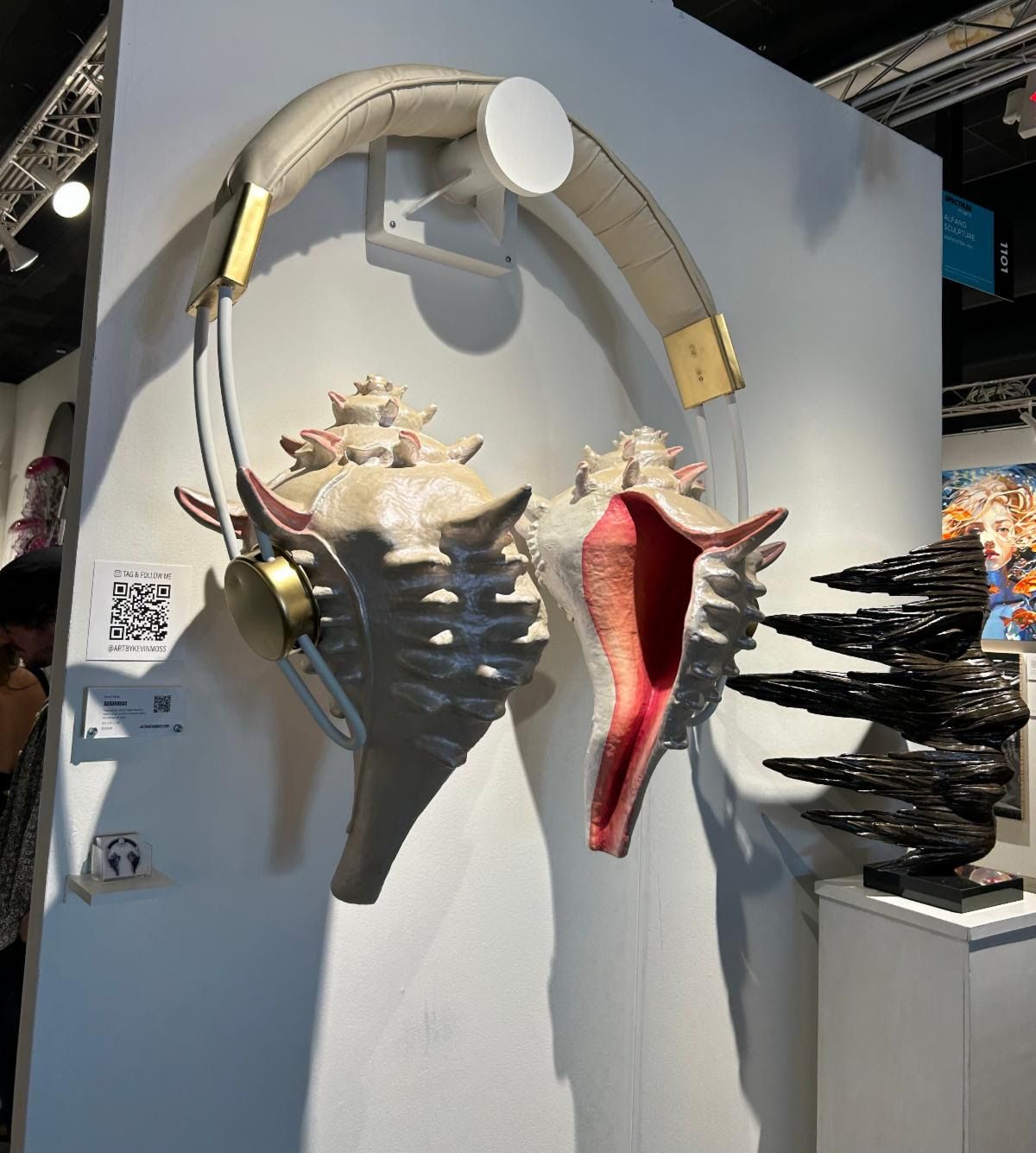 Wall sculpture of giant headphones with spiny conch shells for ear cups, a black carved form on a pedestal beside it.