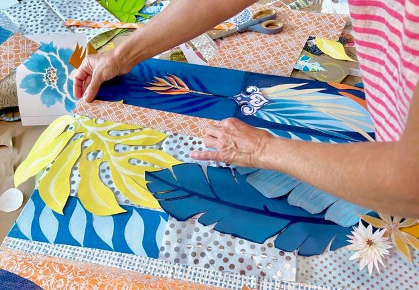 Jennifer's hands placing a yellow monstera cut-out among deep blue banana leaves and silver-dotted papers on a work in progress