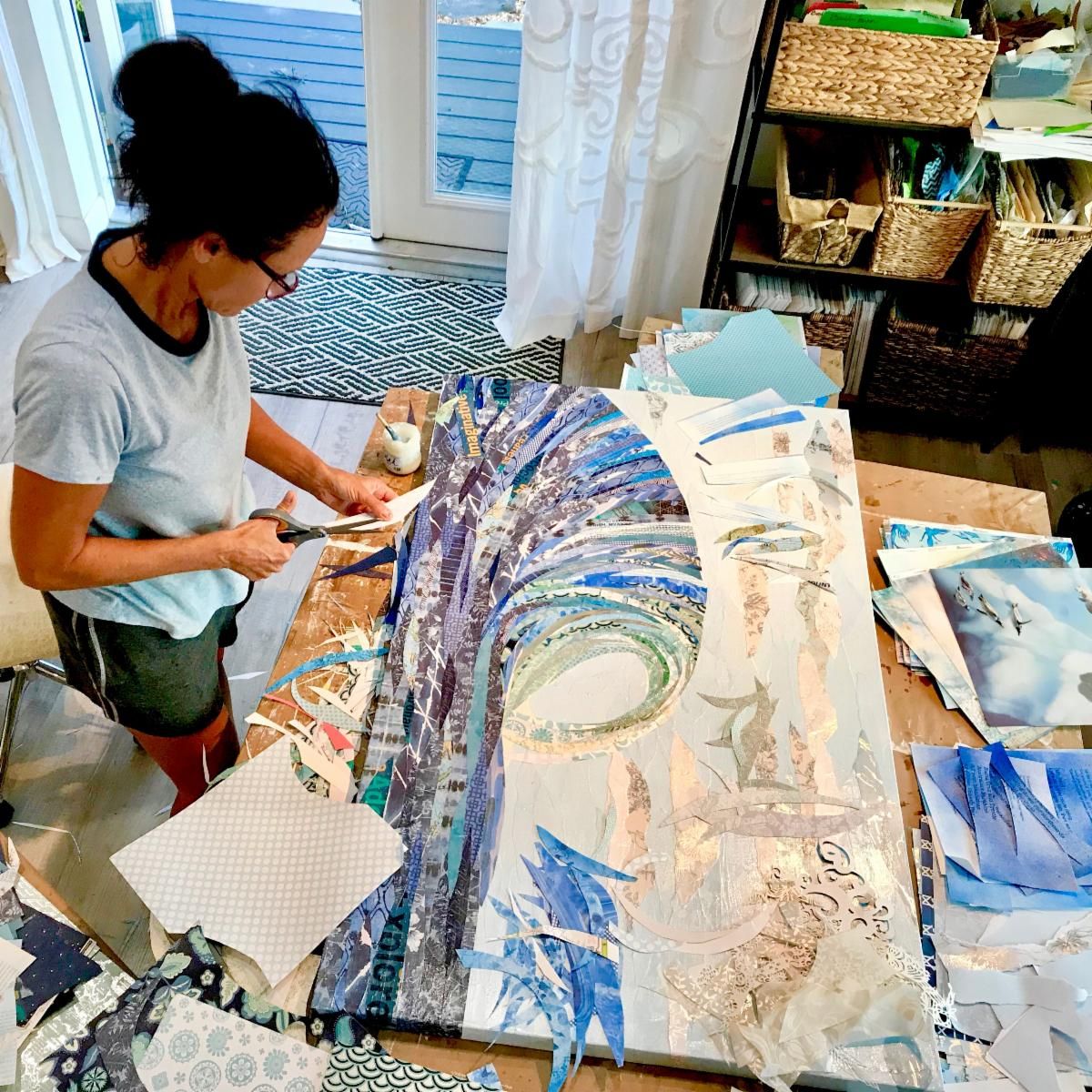 Jennifer stands at her studio table trimming papers for the Big Kahuna wave, baskets of sorted papers shelved behind her