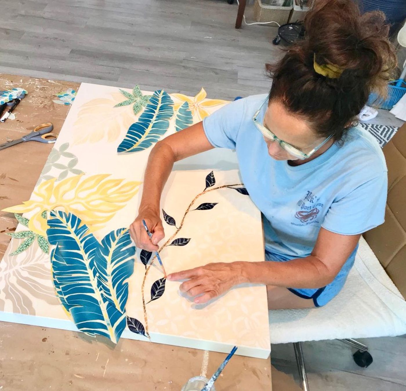 Jennifer paints the navy leaf vine across the Jungle Boogie canvas, golden monstera and teal banana leaves already placed