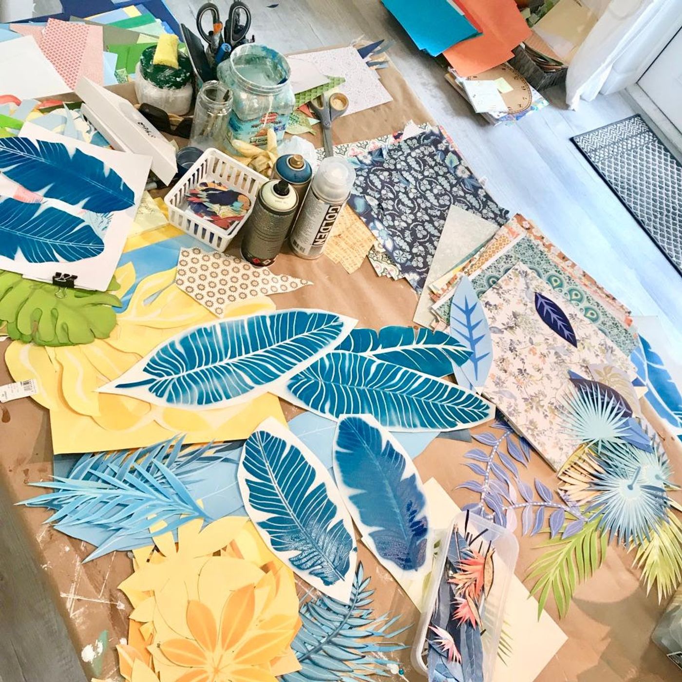 Worktable spread with hand-cut teal banana leaves, palm fronds, stacks of patterned paper, spray cans, and scissors