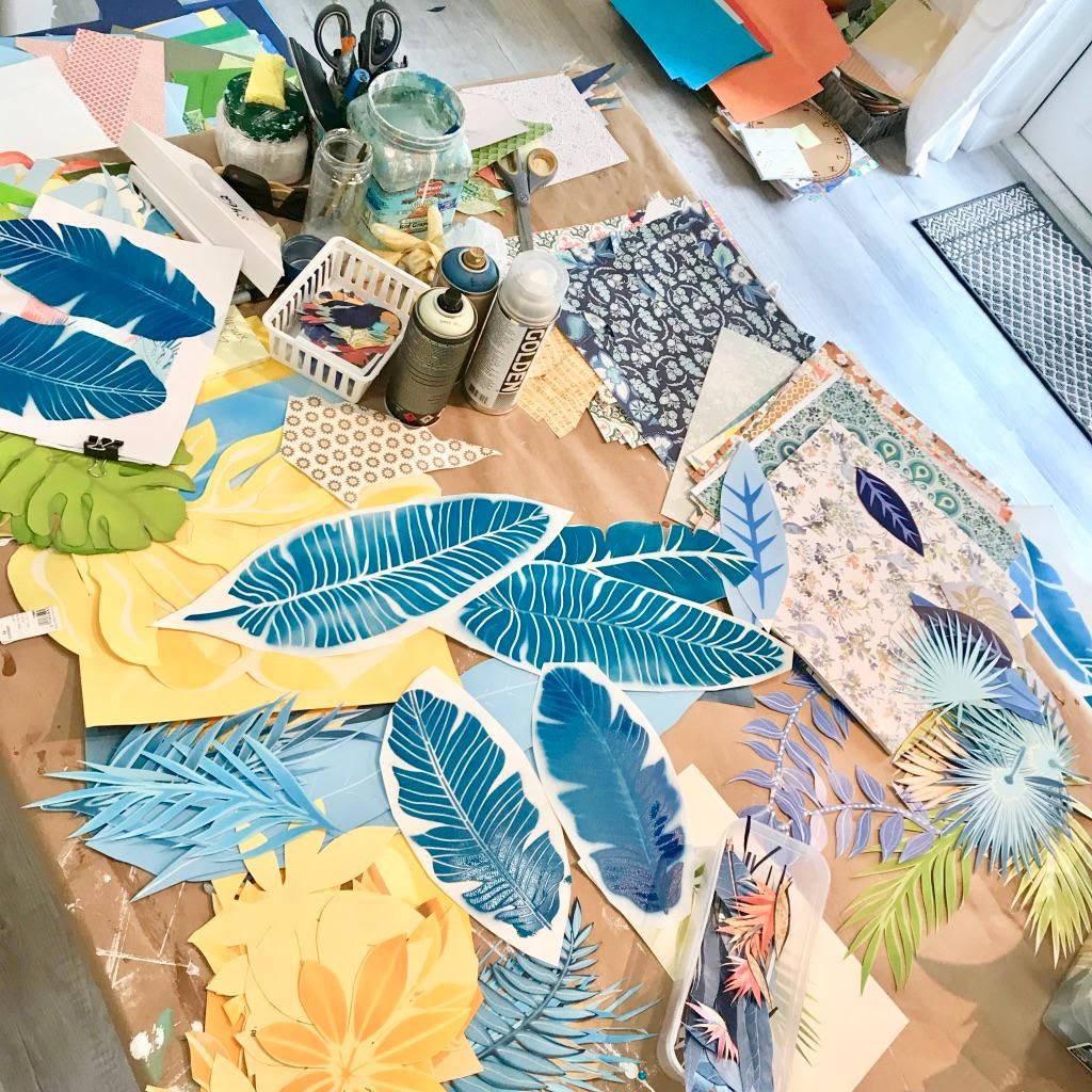 Worktable spread with hand-cut teal banana leaves, palm fronds, stacks of patterned paper, spray cans, and scissors