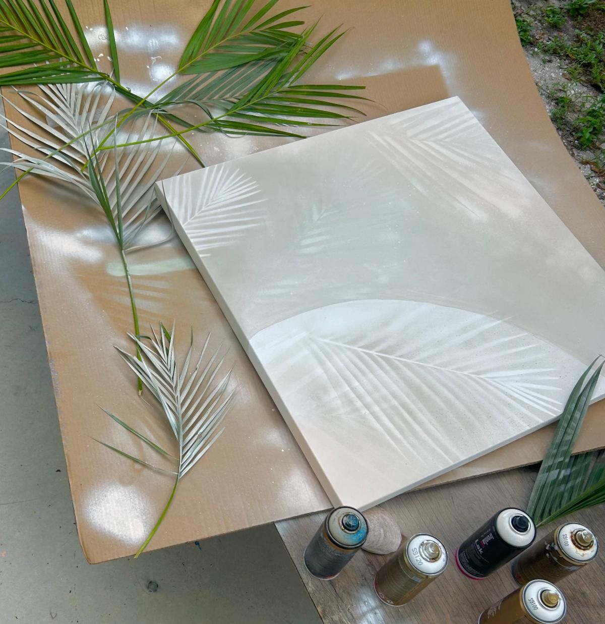 A canvas spray-stenciled with ghostly white palm-frond impressions, silver-painted fronds and spray cans on cardboard outdoors.
