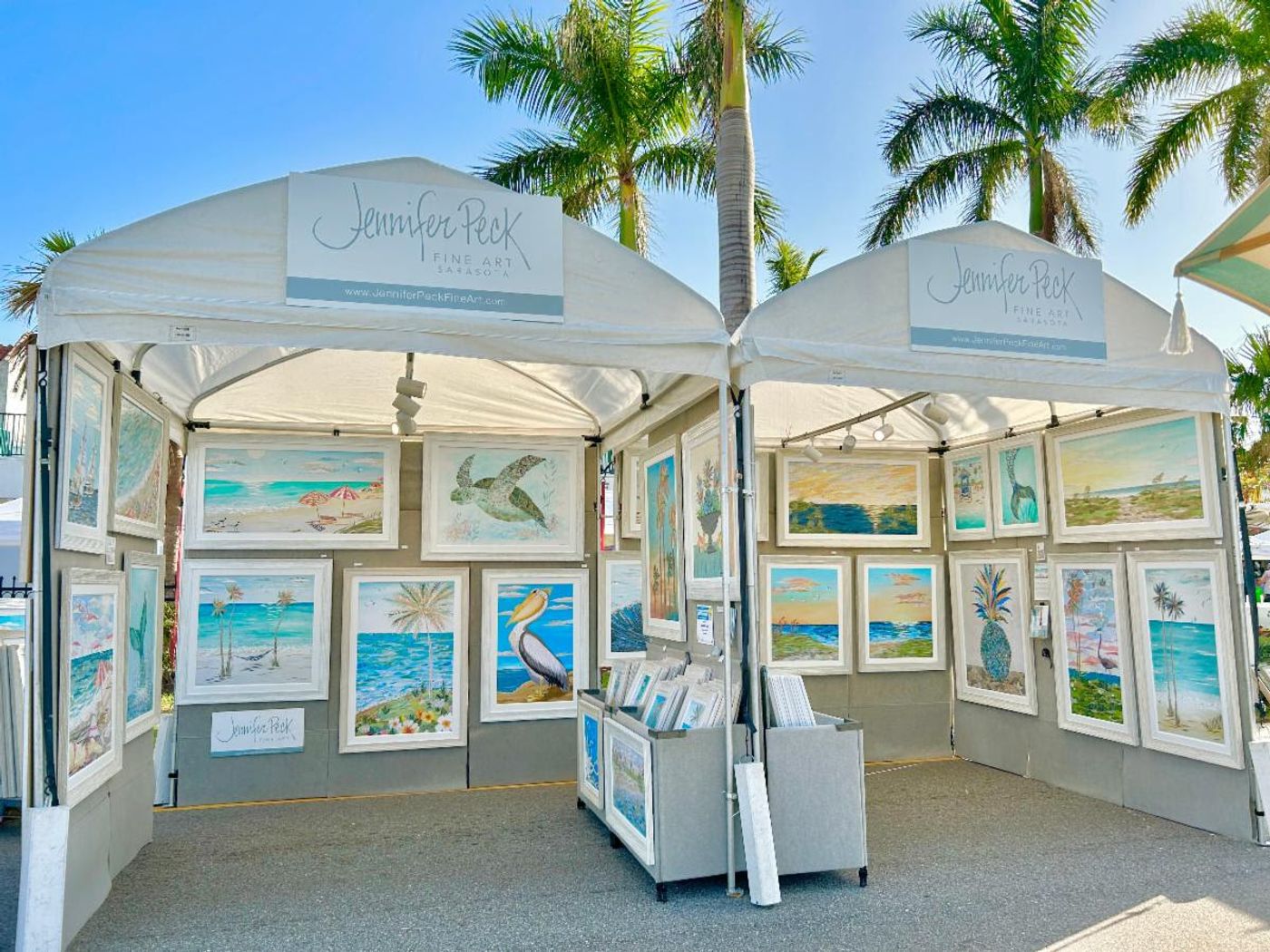Jennifer's double white festival tent under palms, walls lined with framed coastal collages — sea turtle, pelican, beach scenes