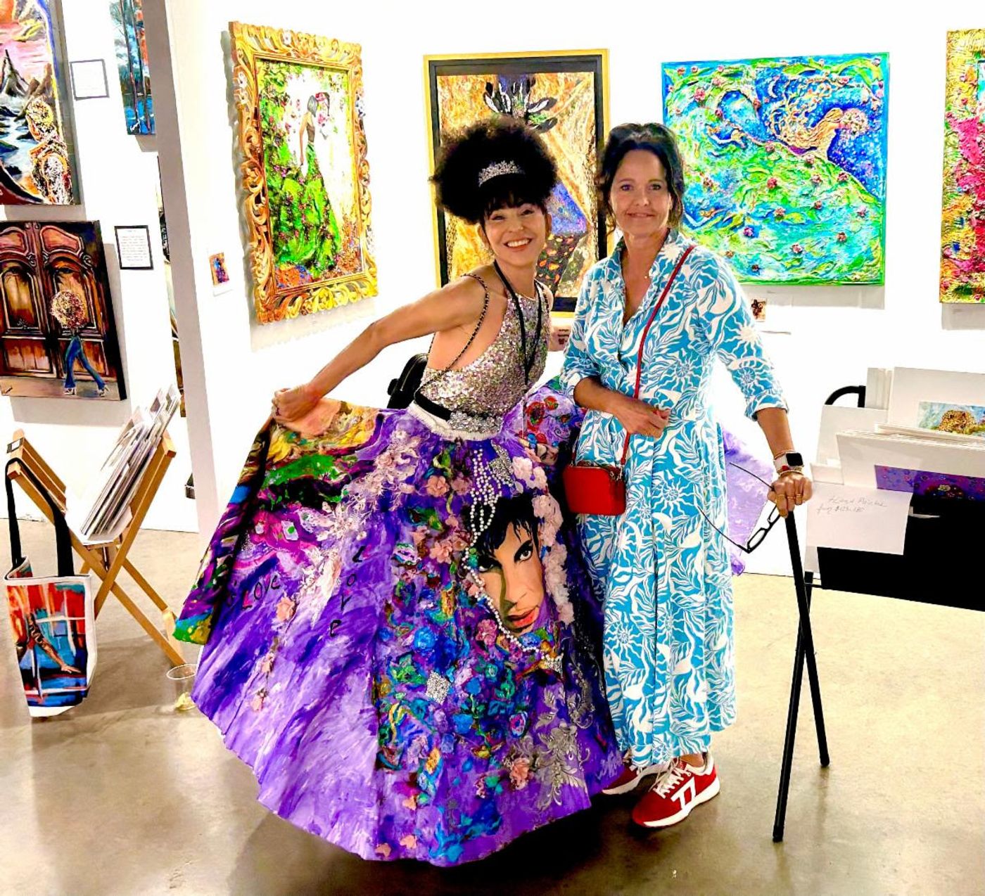 Jennifer in a blue-and-white dress poses with a woman in a purple Prince-painted ballgown at a Miami art fair booth.
