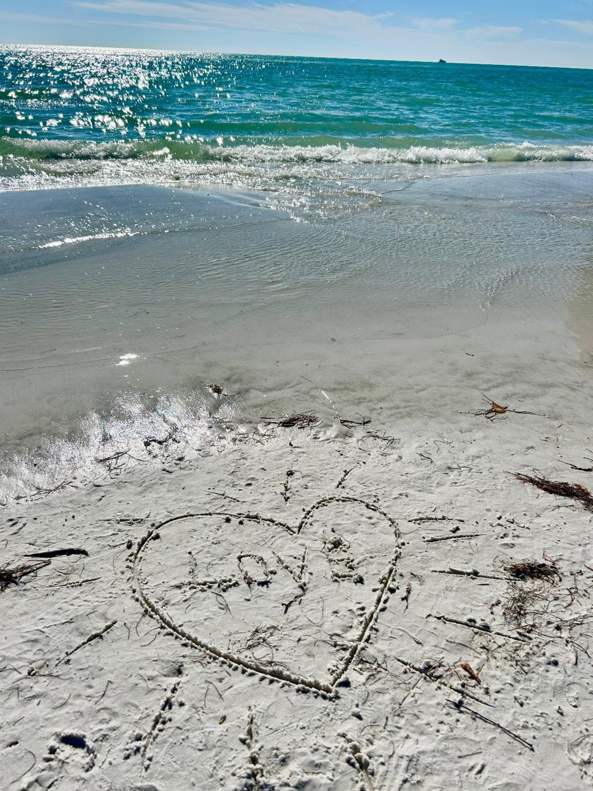 A heart traced in the sand at the water's edge with 'Love' written inside, sparkling turquoise Gulf beyond